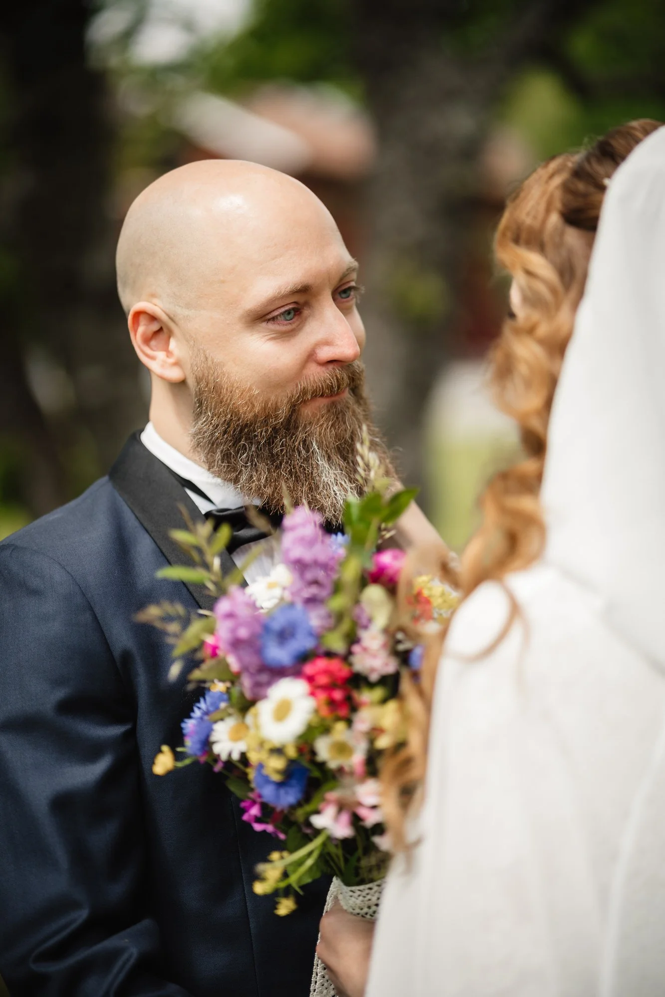 A man with a beard in a tuxedo holding colorful flowers, standing face to face with a woman dressed in white, both at an outdoor event.