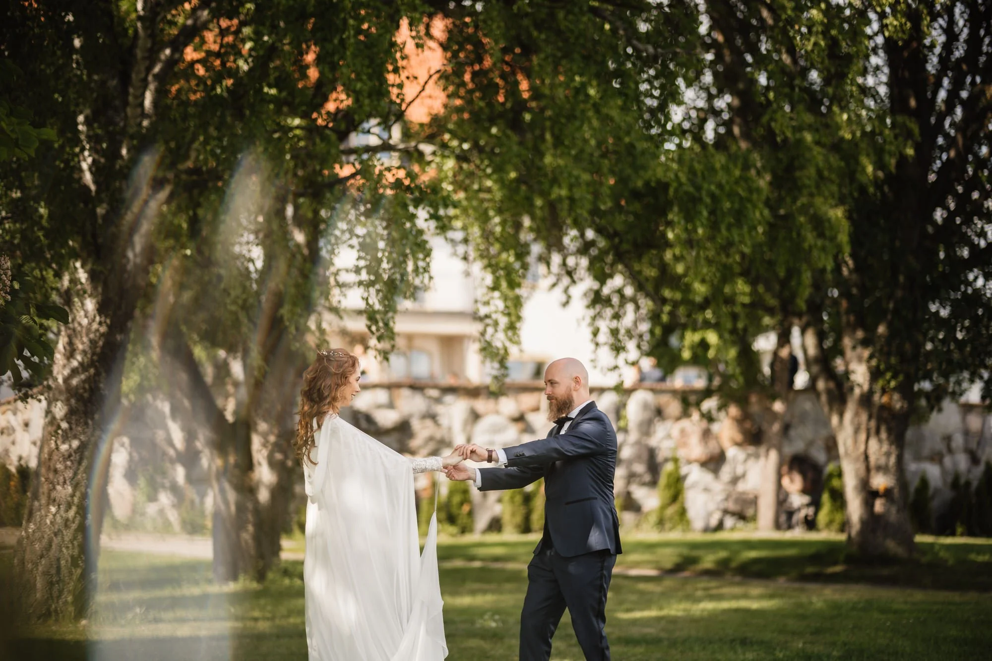 En brud och en brudgum står under ett träd och håller varandra i händerna i en park, troligen under en bröllopsceremoni på eftermiddagen.
