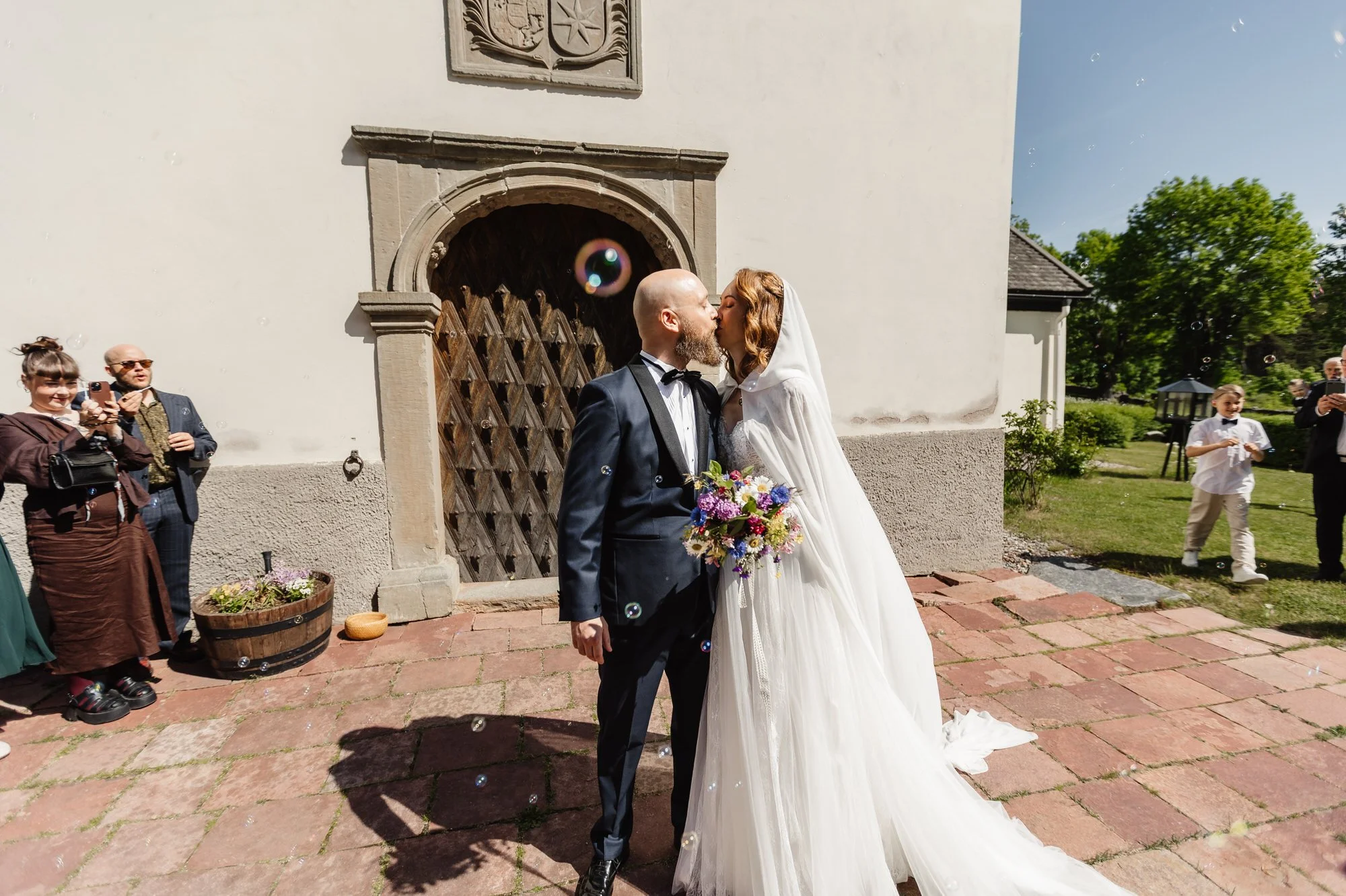 Nygifta par kysser utanför en vit kyrka, med gäster som tittar och tar bilder.