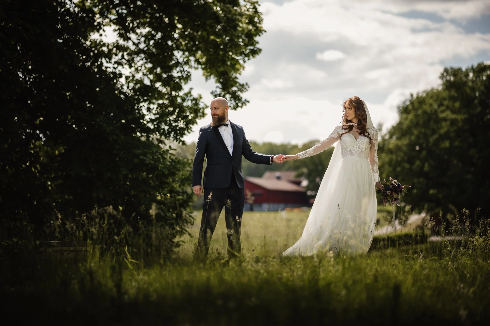 En brud och en brudgum som håller varandra i handen ute i en grön äng under en molnig himmel.