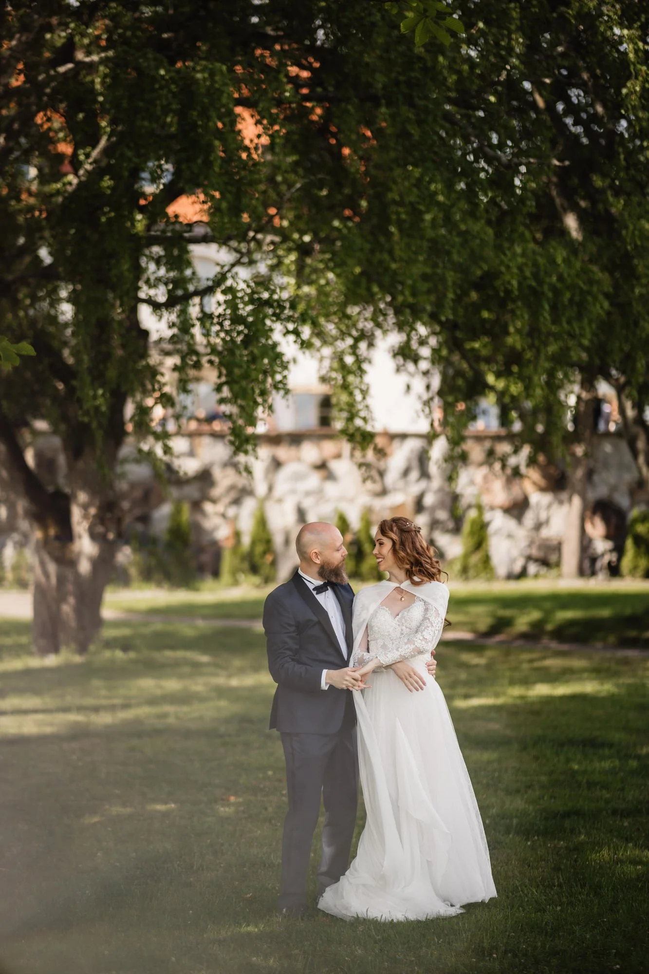 En man och en kvinna klädda i bröllopskläder står tillsammans i en park med grönt gräs och stora träd. Mannen är klädd i svart kostym och kvinna i vit brudklänning. De ser på varandra och håller varandras händer.