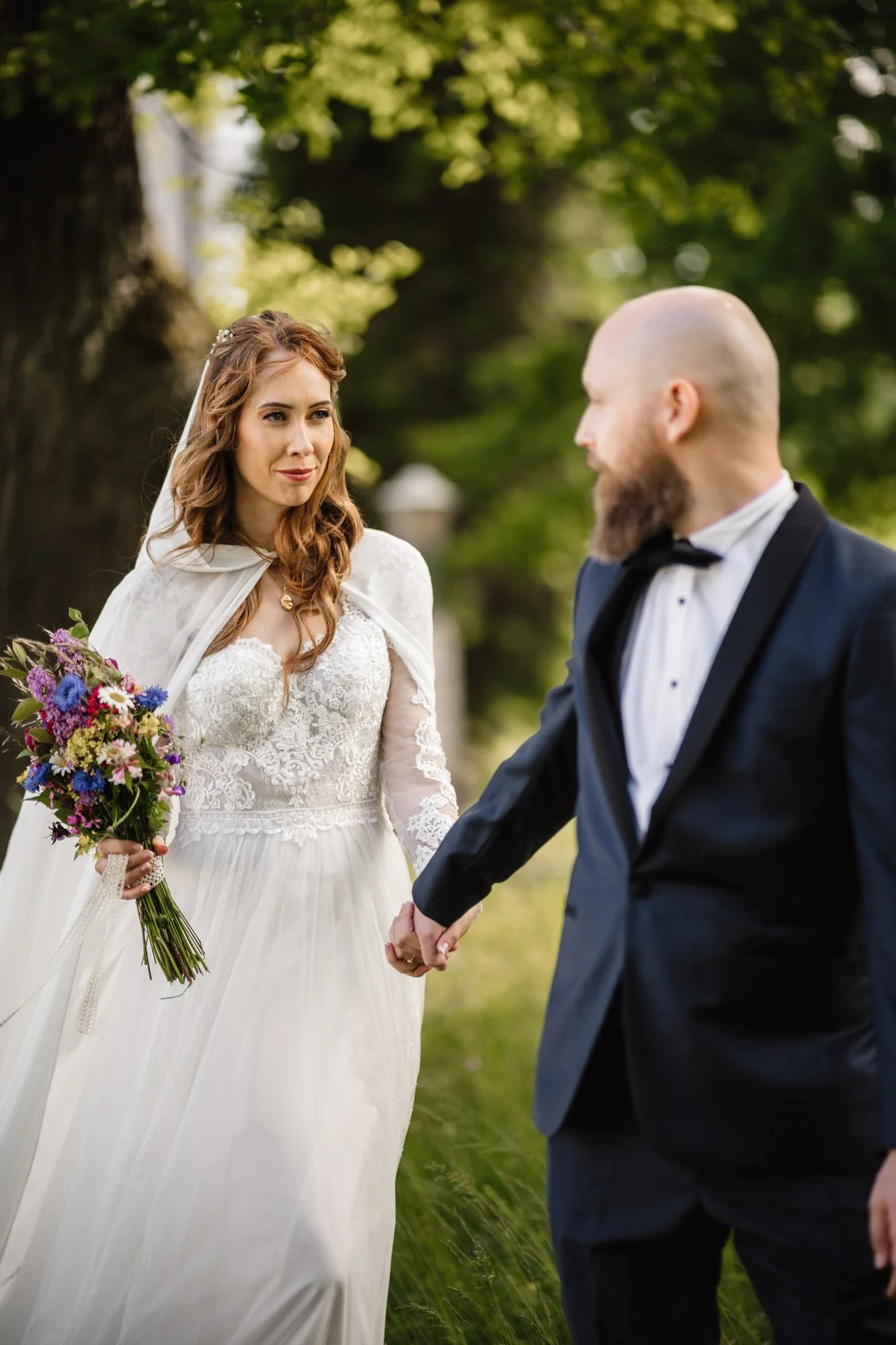En brud och en brudgömma håller hand utomhus under ett träd, bruden bär en vit brudklänning och håller en blombukett, de ser varandra i ögonen.
