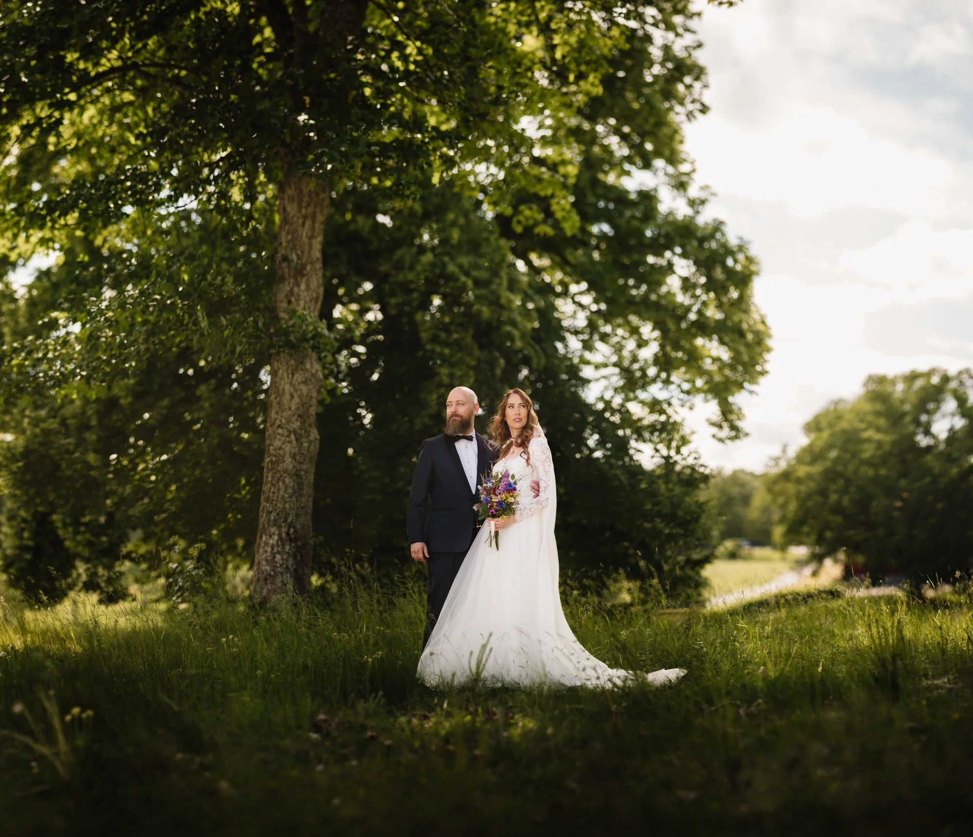 I bilden syns ett brudpar, en man i svart tuxedo och en kvinna i en vit brudklädsel, stående i en grön park med stora träd bakom dem. De håller varandra och ser ut att vara på ett bröllop.