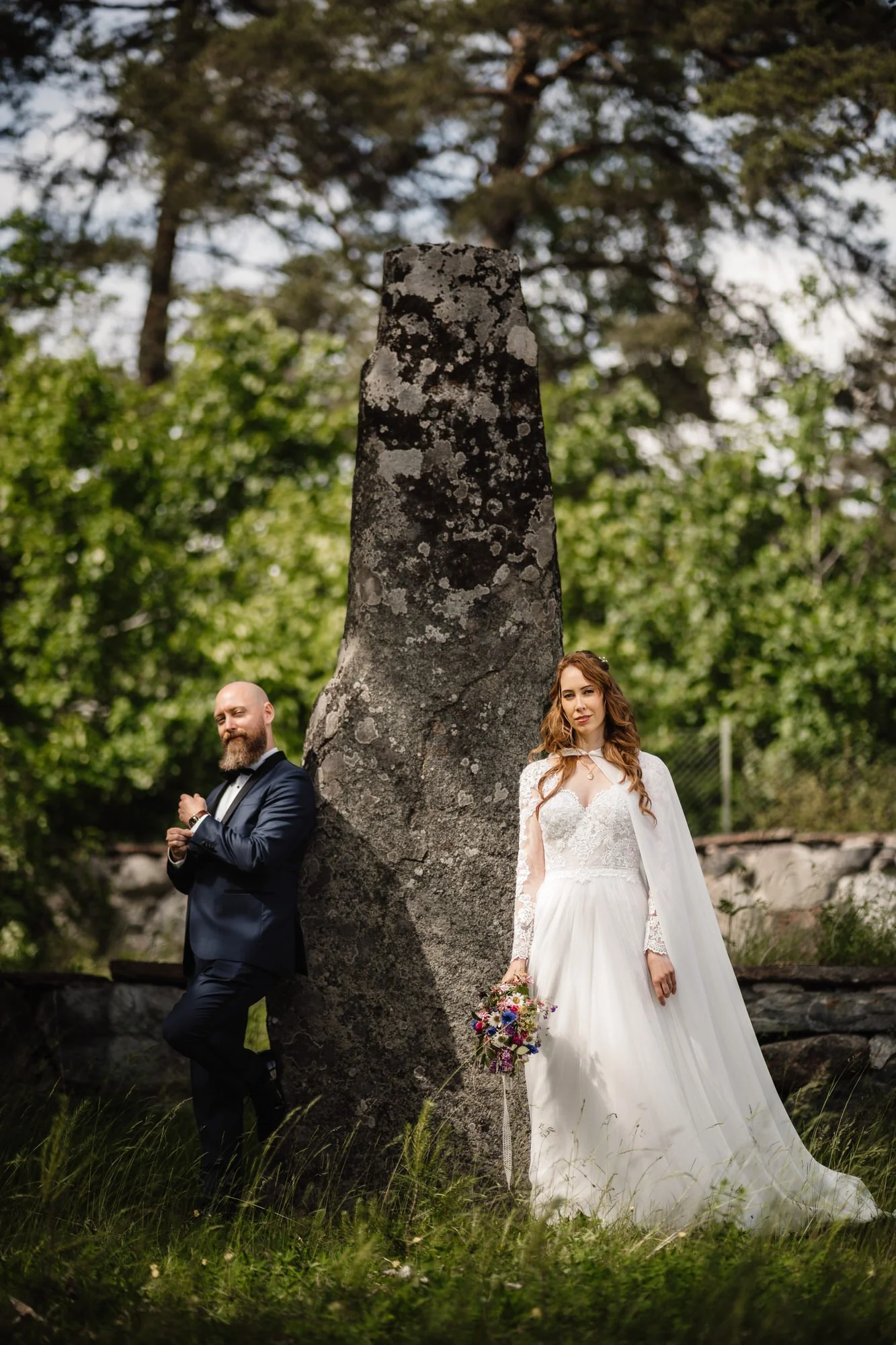 En nygift man och kvinna står på varsin sida av ett stort, mossigt träd i en grön park. Mannen är klädd i en mörk kostym och kvinnan i en vit brudklänning och håller en bukett blommor.