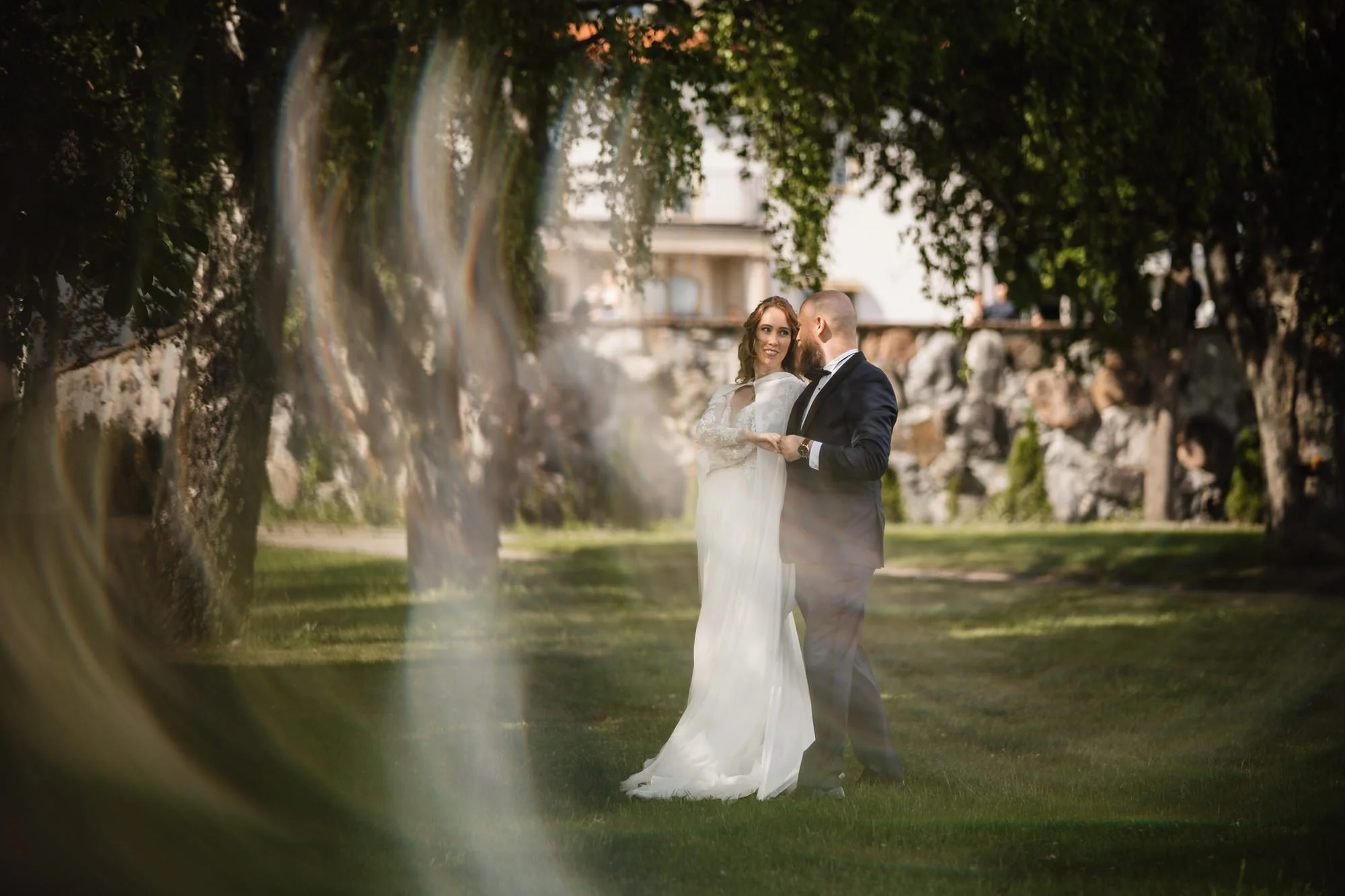 En nygift brud och brudgum som håller varandra i handen utomhus, omgivna av gröna träd och en byggnad i bakgrunden, troligen under en fotografering.