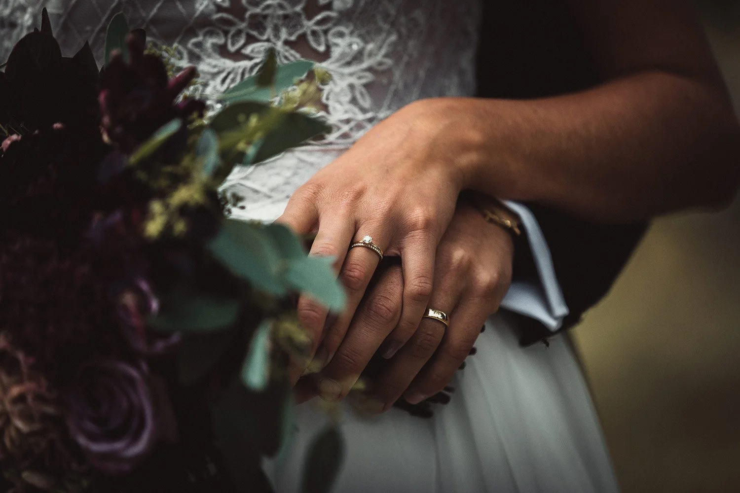 Närbild på brudparets händer och vigselringar med bukett och spetsdetaljer under ett elegant bröllop.
