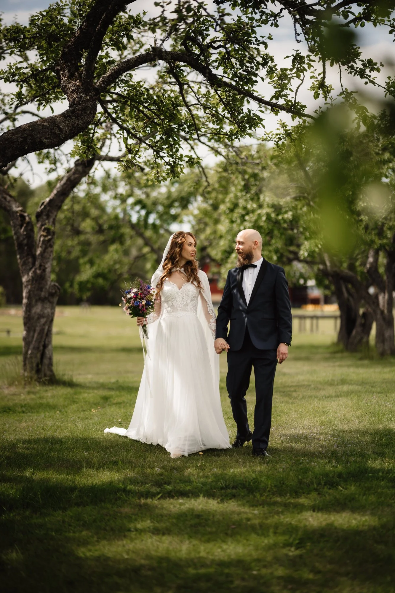 En ung kvinna i en vit brudklänning och en man i svart tuxedo går hand i hand i en grön park med träd, de ser varandra i ögonen och ler.