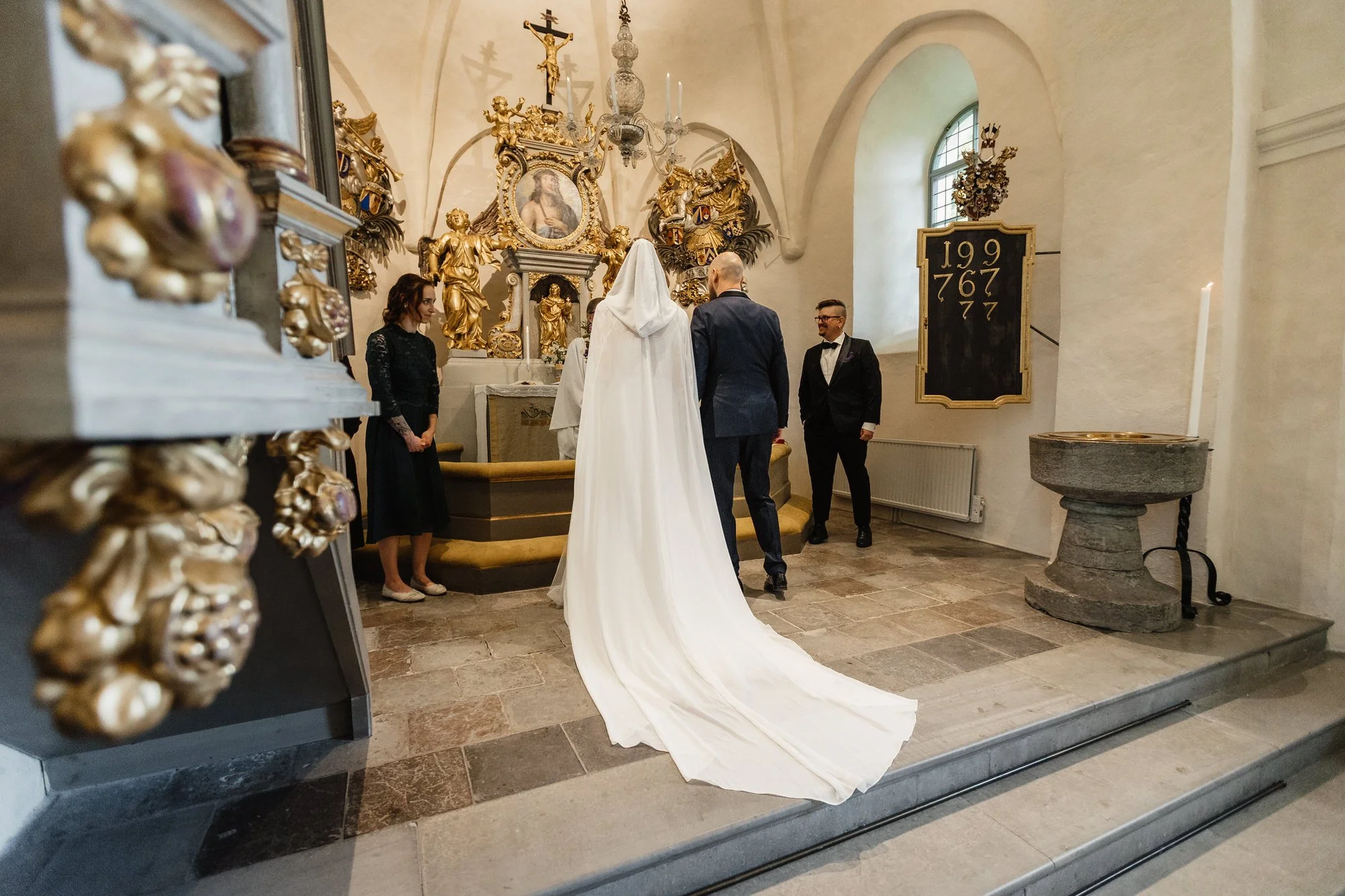 Vackert bröllopsceremoni i en kyrka med präster och brud i vit brudklänning. Gulddekor och religiösa konstverk i bakgrunden.