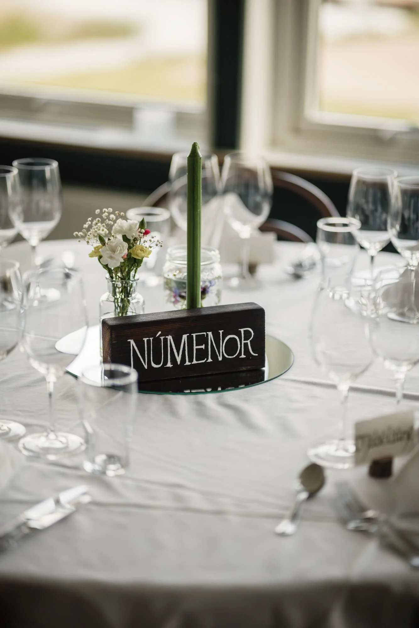 Bord dukat för ett bröllop eller fest med ett blomarrangemang och ett bordsskylt med texten "NÚMENOR". Det finns flera tomma vinglas och bestick runt bordet.