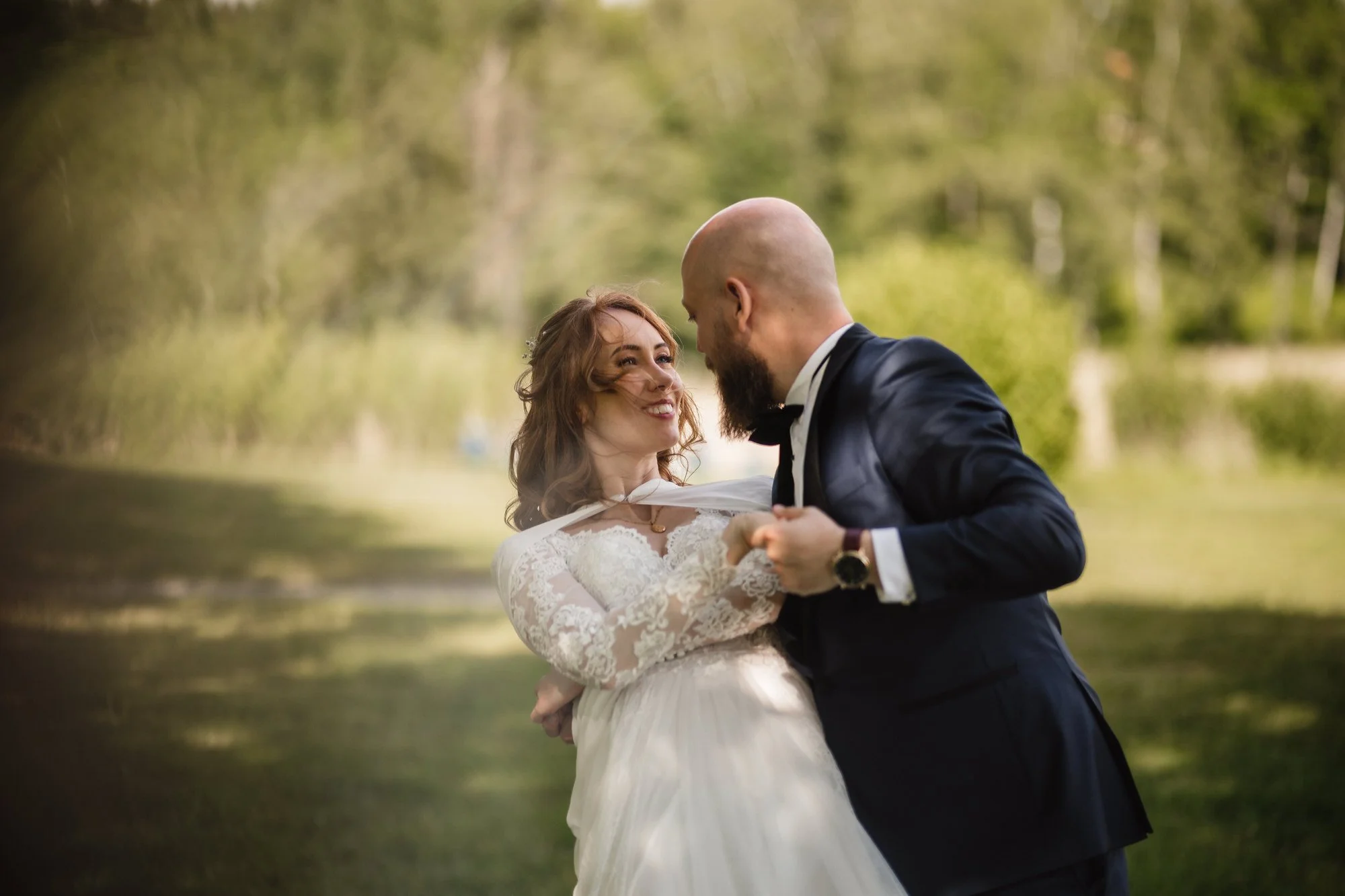 En brud och en bröllopsk groom dansar utomhus vid en sjö, omgivna av gröna träd, ler och tittar hungrigt på varandra.