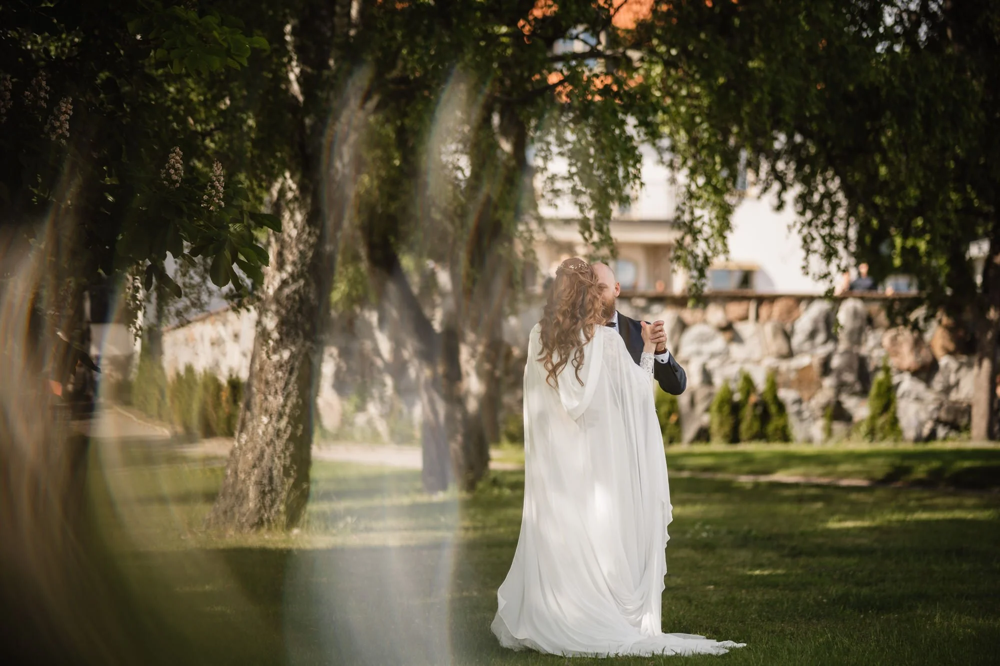 En nygift brud och brudgum dansar tillsammans utomhus i en park, omgivna av gröna träd och gräsmatta, med en byggnad synlig i bakgrunden på kvällen.