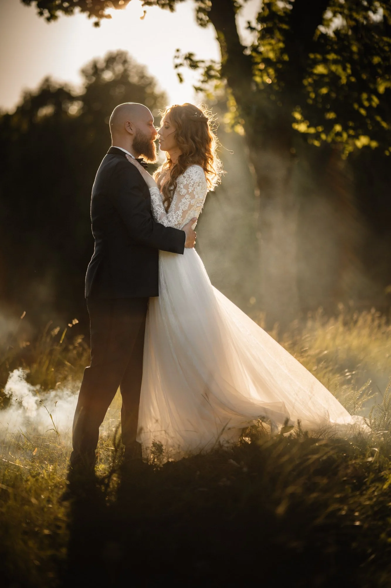 En nygift brud och brudgumo står nära varandra i ett solnedgångslandskap. Bruden bär en vit, romantisk brudklänning med långärm och spetsdetaljer, medan brudgumen är klädd i en svart tuxedo. Solens ljus skiner igenom trädens löv i bakgrunden, vilket 