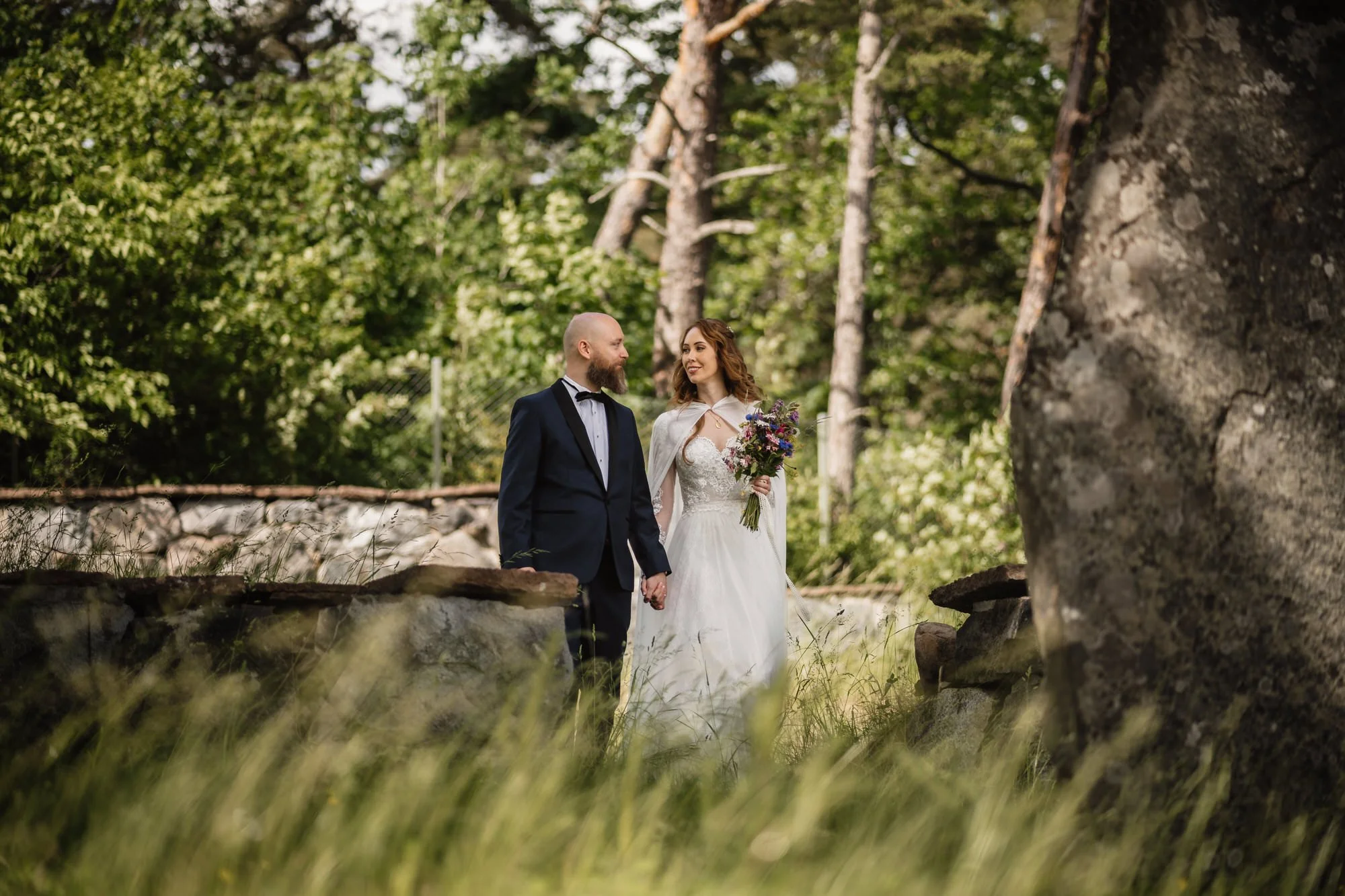 En brud och en brudgum står tillsammans i en skog, håller handen och tittar på varandra, där bruden håller en bukett blommor.