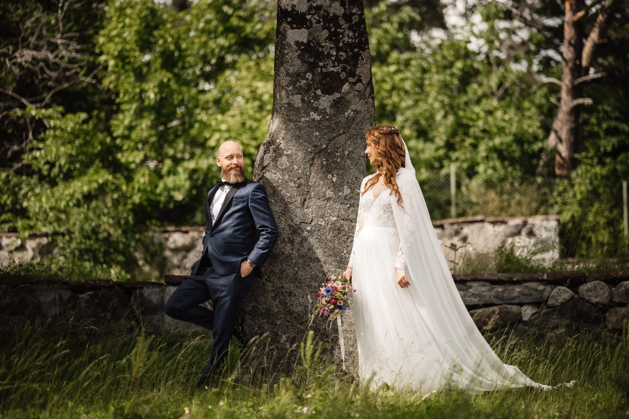 En man och en kvinna i bröllopskläder står under en stor ek i en grön park, kvinna håller en bukett blommor, de tittar på varandra.