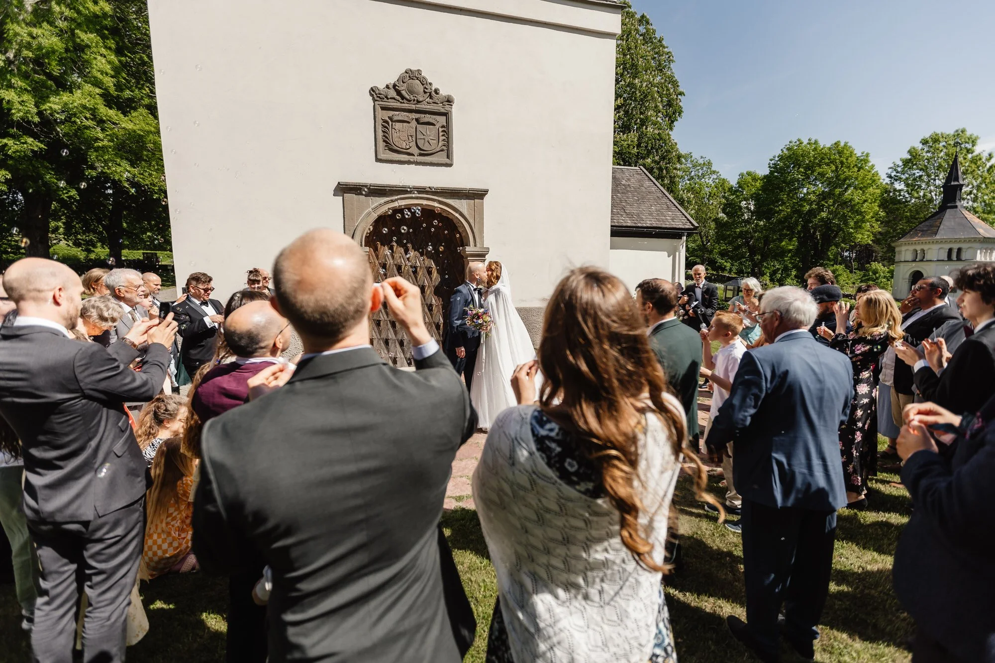 En bröllopsceremoni med ett brudpar framför en vit byggnad, omgiven av gäster, under en solig dag med gröna träd i bakgrunden.