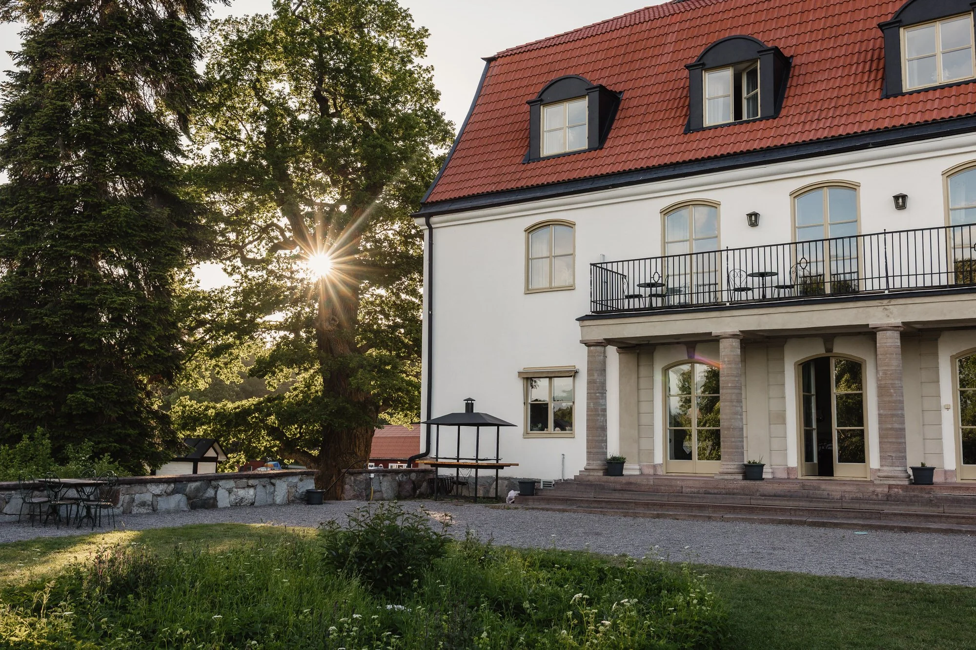 Ett stort vitt hus med blå detaljer, en balkong med svart räcke, träd och solnedgång i bakgrunden.