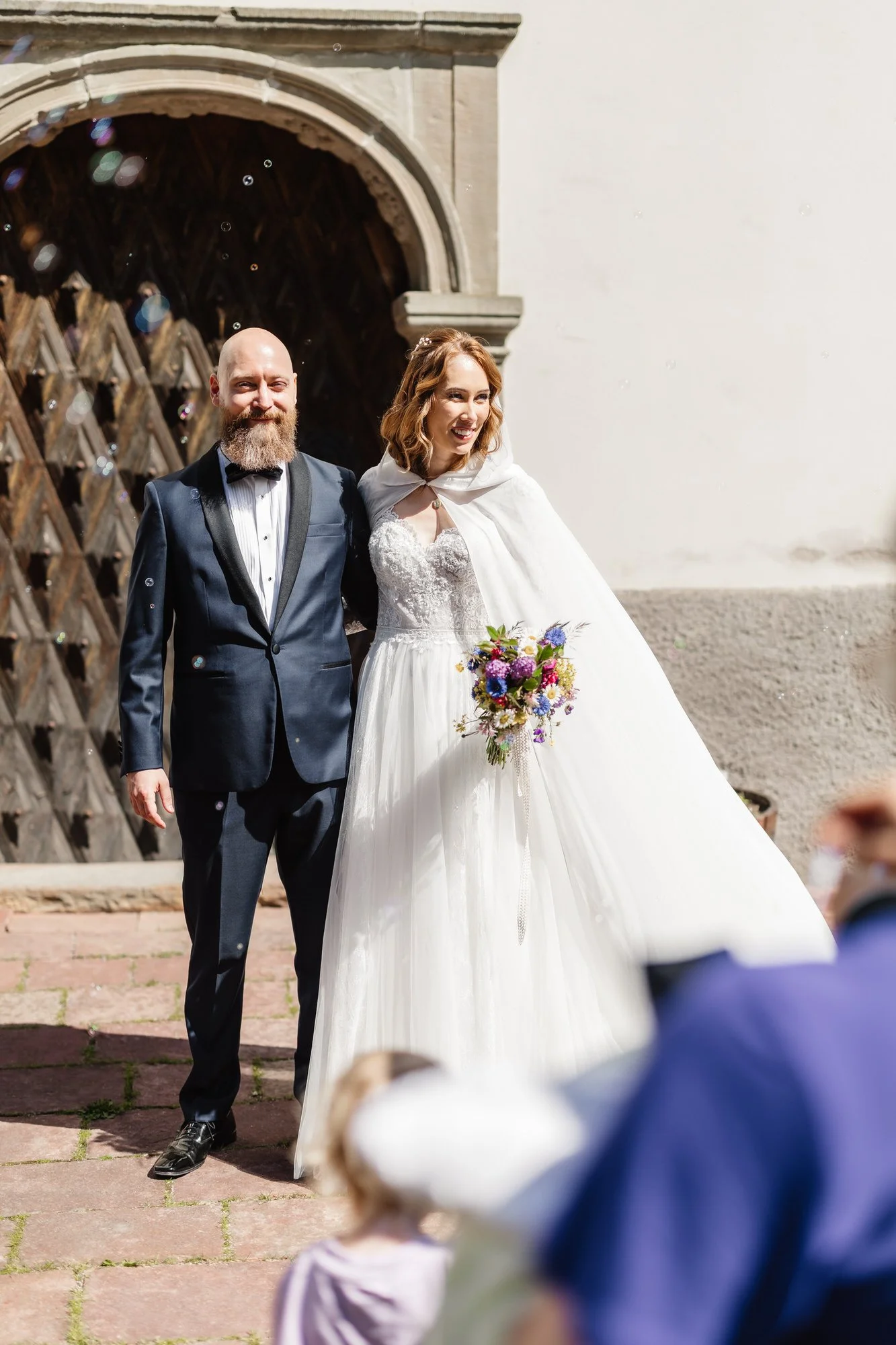 En man och en kvinna på bröllop står tillsammans utomhus utanför en äldre byggnad, mannen bär en mörk kostym och kvinna en vit brudklänning med ett färgglatt blomsterarrangemang, de ler och ser glada ut.