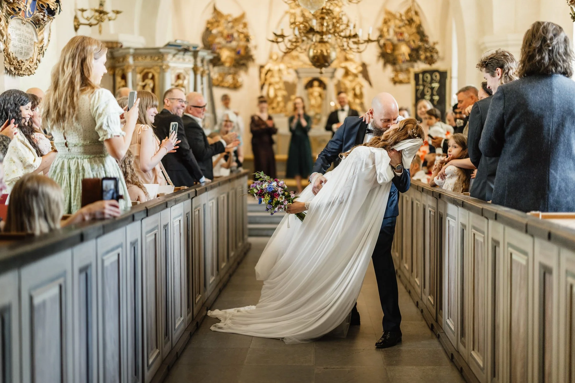 En brud och en gäst kysser varandra i en kyrka under ett bröllop, omgivna av gäster som tar bilder och tittar.