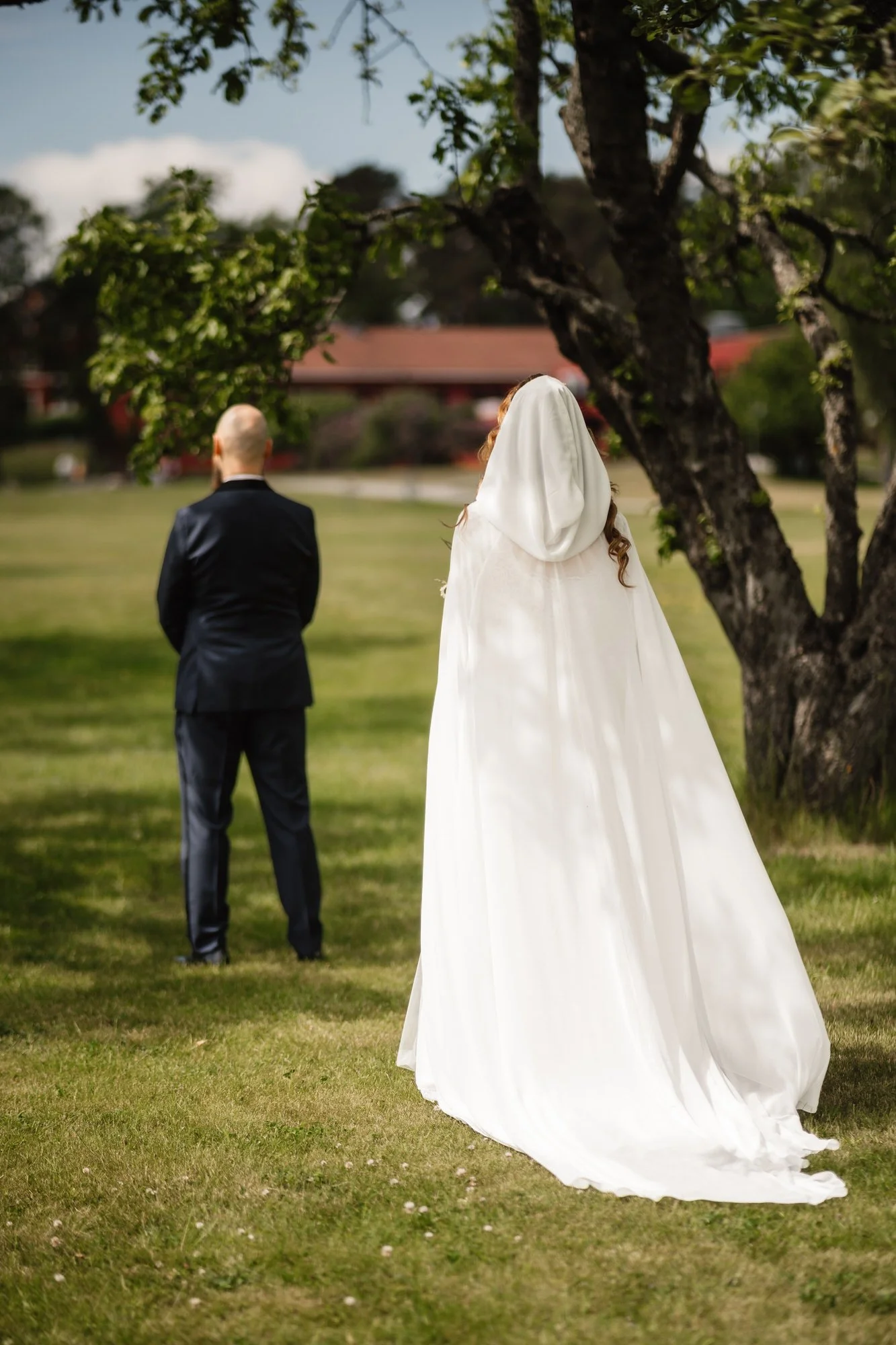En brud och en brudgum ses utomhus, med brudens rygg vänd mot kameran, klädd i en vit brudklänning med släp, och brudgummen i en mörk kostym, under ett träd på en gräsmatta.