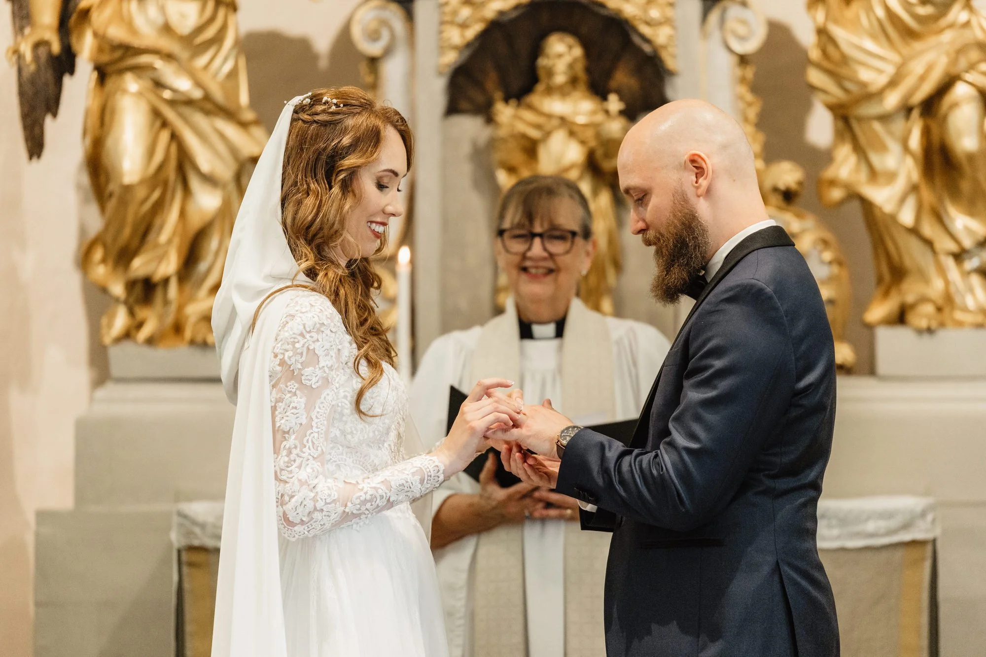 En brud och en brudgum som håller händer under deras vigselceremoni i en kyrka, med en präst som bevittnar, och en bakgrund av gyllene dekorationer.