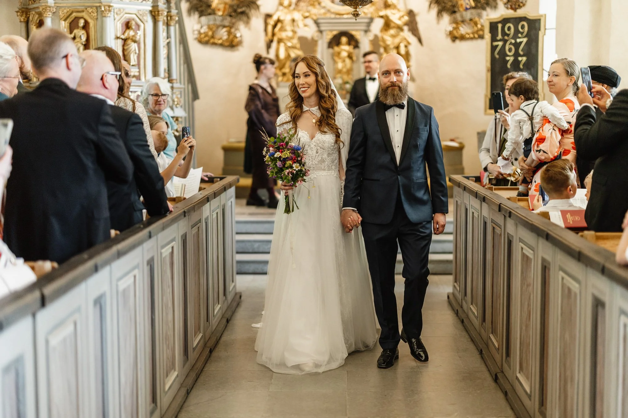En brud och en brudgum som håller varandra i handen i kyrkbänkarna under ett bröllop, omgiven av gäster som fotograferar och ser på, i en kyrka med eleganta gulddekorationer.