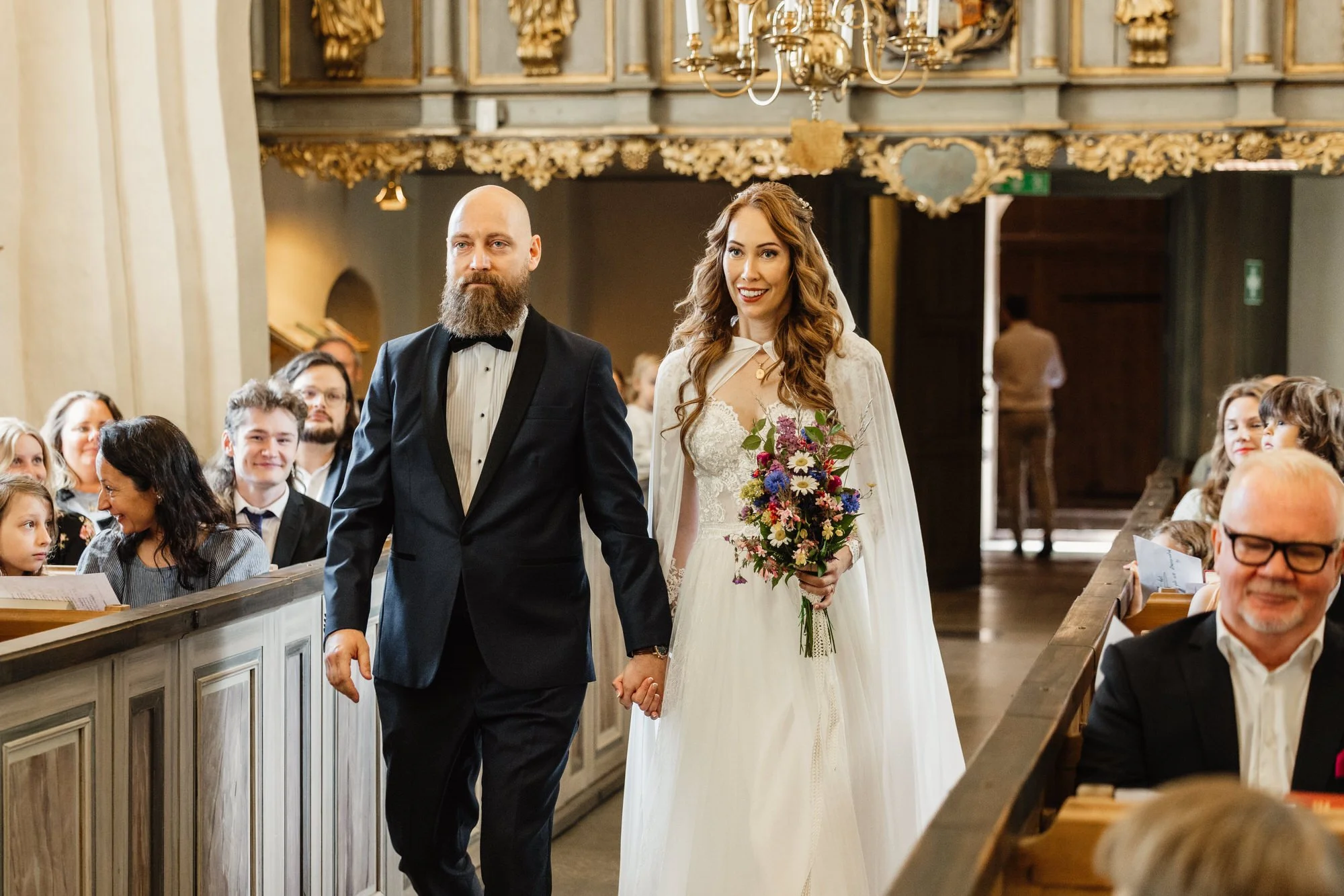 En brud och en brudgum går nerför gången i en kyrka, den kvinnliga bruden håller en stor bukett blommor och ler, medan den manliga brudgummen har en allvarlig min. Personerna i bakgrunden är sitta i bänkar i kyrkan, omgivna av vackra gulddekorerade v
