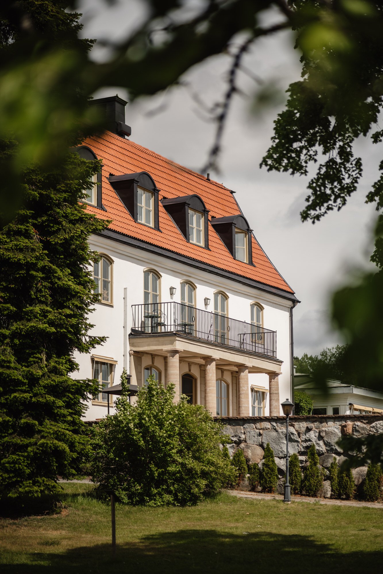 Tammsviks herrgård med ett rött tegeltak och flera fönster, omgiven av gröna träd och blomster. En balkong med stol och bord syns på andra våningen.