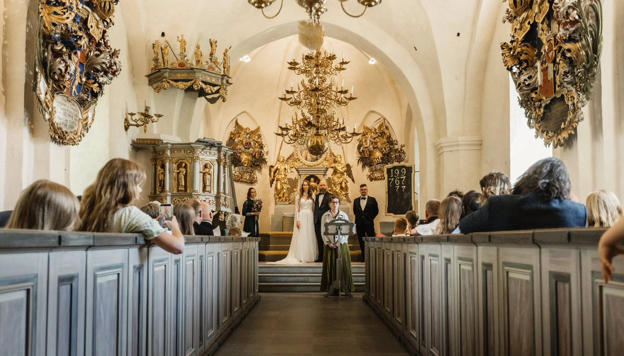 Bröllopsceremoni i en klassisk kyrka med höga tunnvalv, guldfärgade dekorativa skulpturer och ett stort kristallkronlampor. Brud och brudgummi står framför altaret, omgivna av gäster.