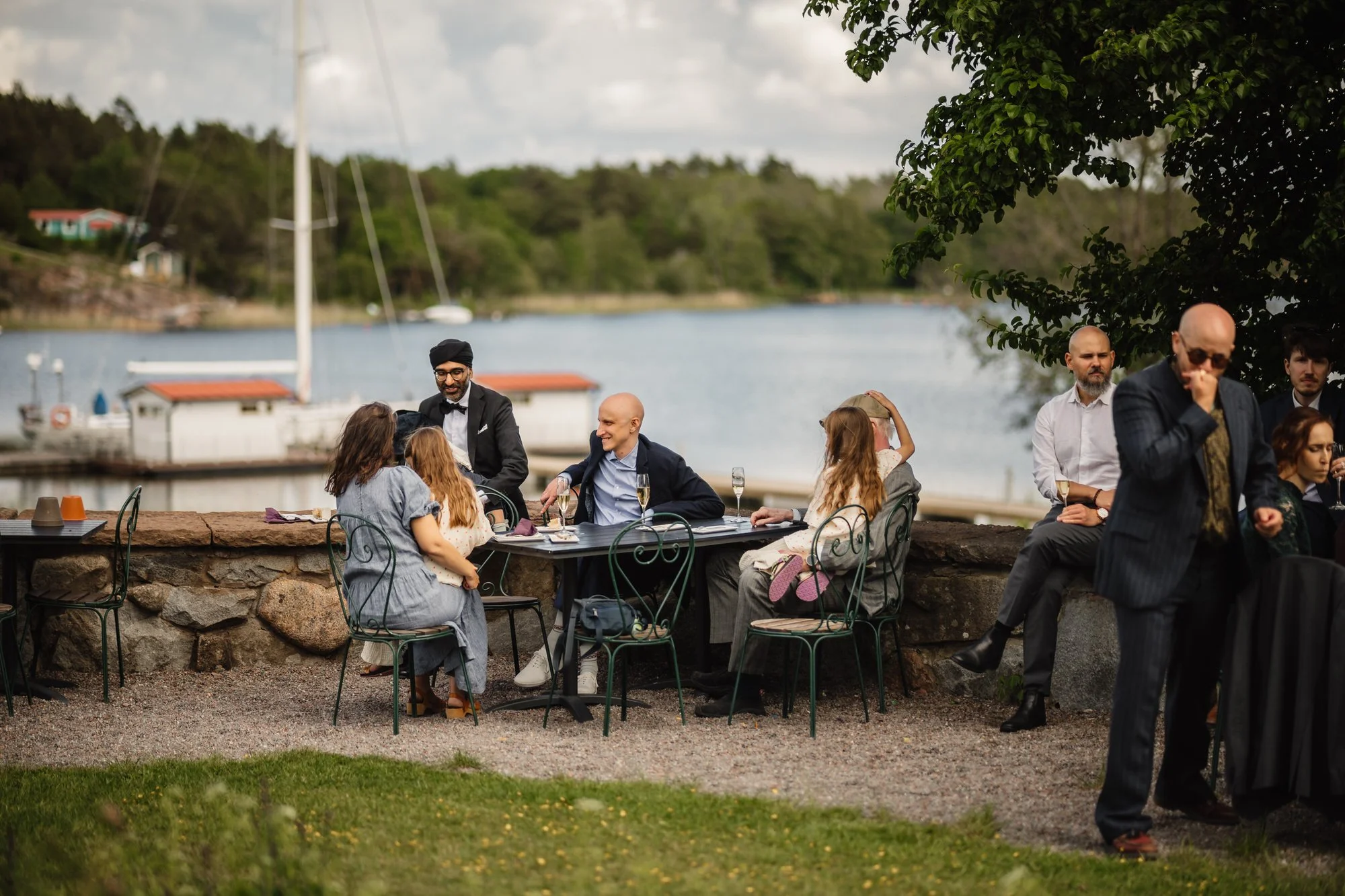 Grupp av människor i formell klädsel vid ett utomhusmöte vid vattnet som inkluderar middagar och samtal, med en båthamn och grönskande natur i bakgrunden.