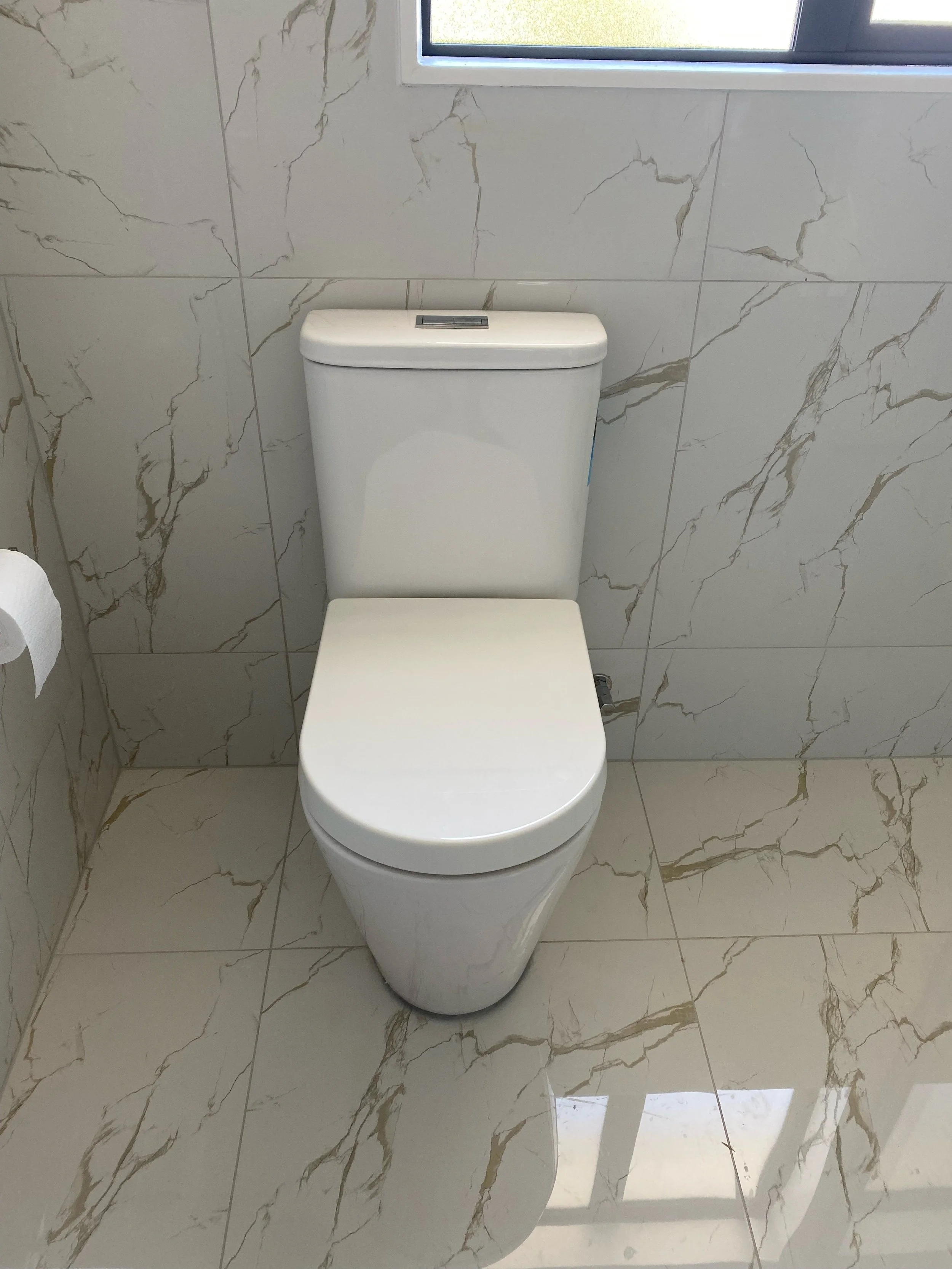 White modern toilet in a bathroom with marble-patterned tiles on walls and floor, and a small window above the toilet.