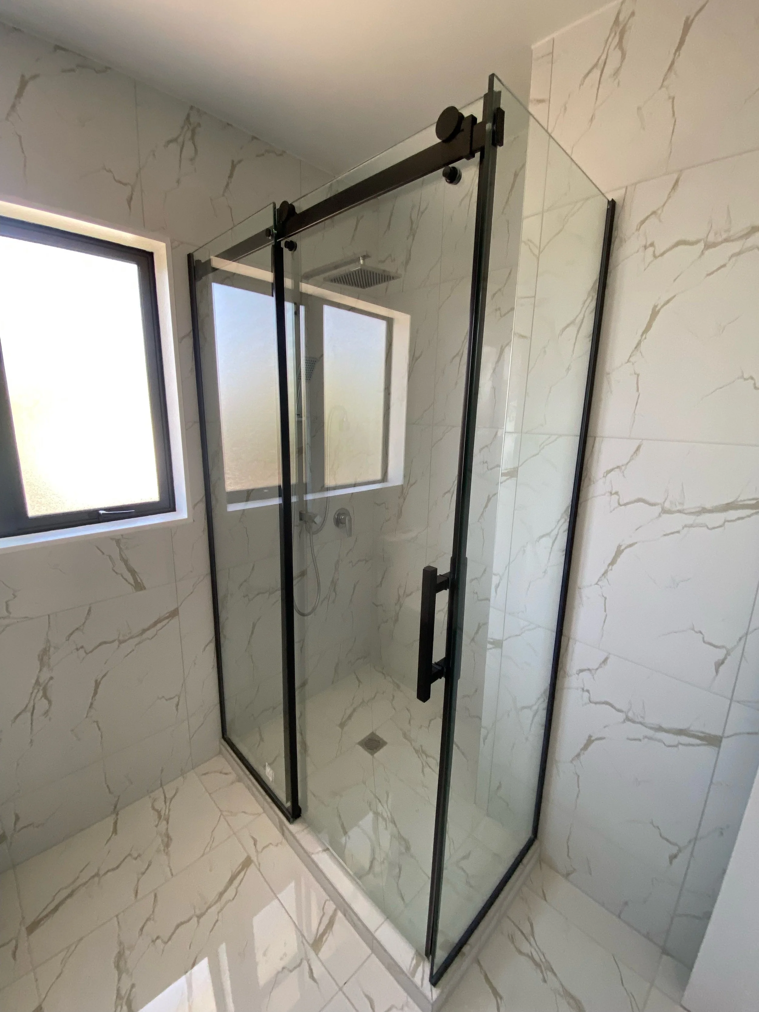 Modern walk-in shower with clear glass enclosure, black frame, marble wall and floor tiles, and a window with frosted glass.