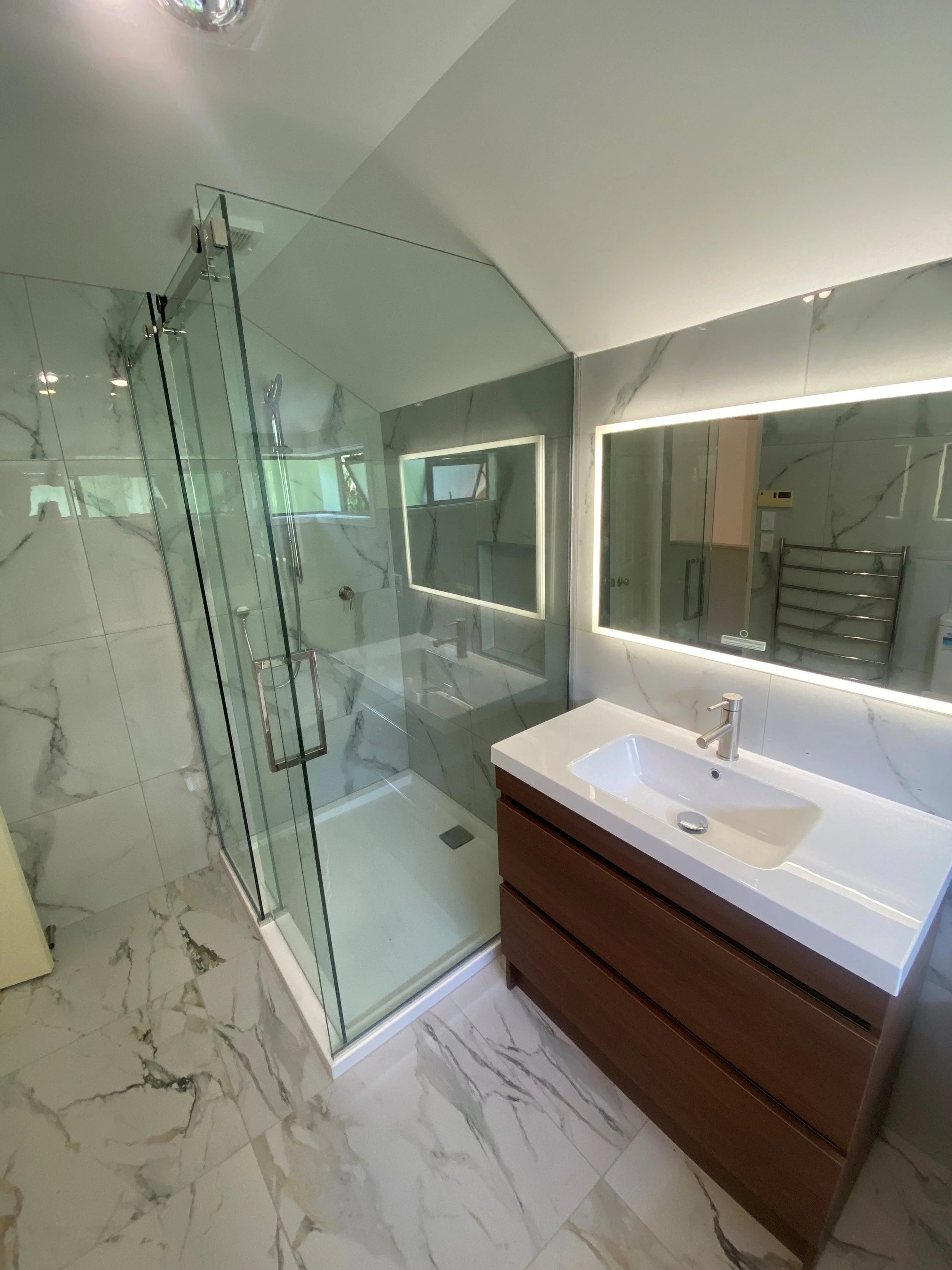 Modern bathroom with marble tile floors, a glass shower enclosure, a rectangular white sink on a wooden vanity, and illuminated rectangular mirrors.