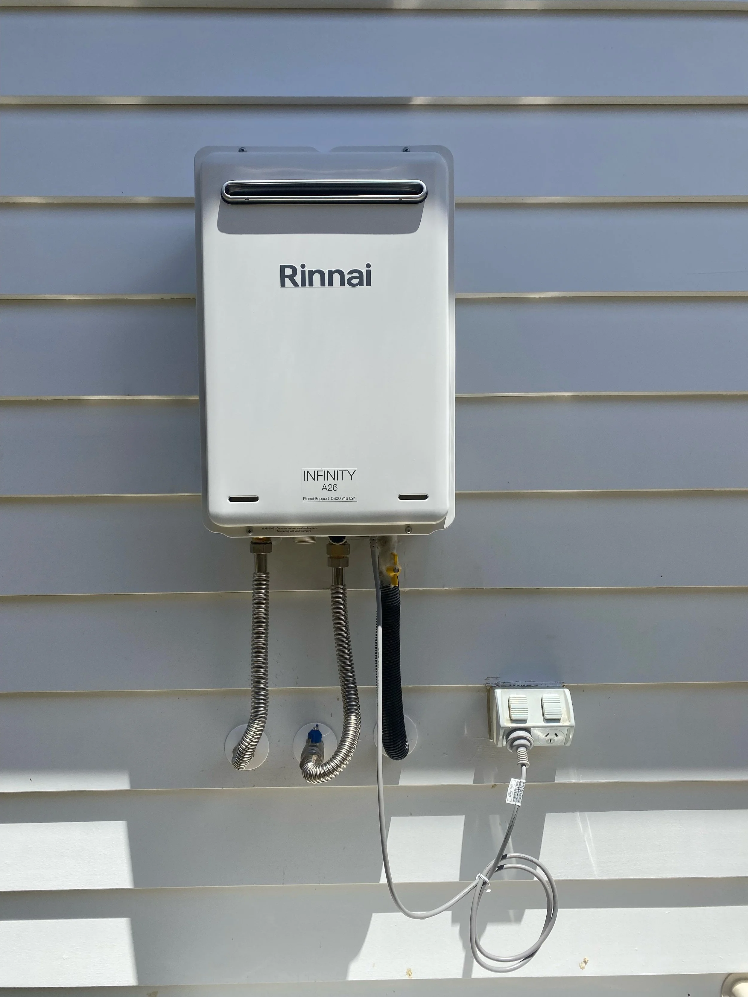A white Rinnai Infinity tankless water heater mounted on a white exterior wall, with three pipes connected at the bottom and an electrical outlet with a cord plugged in.