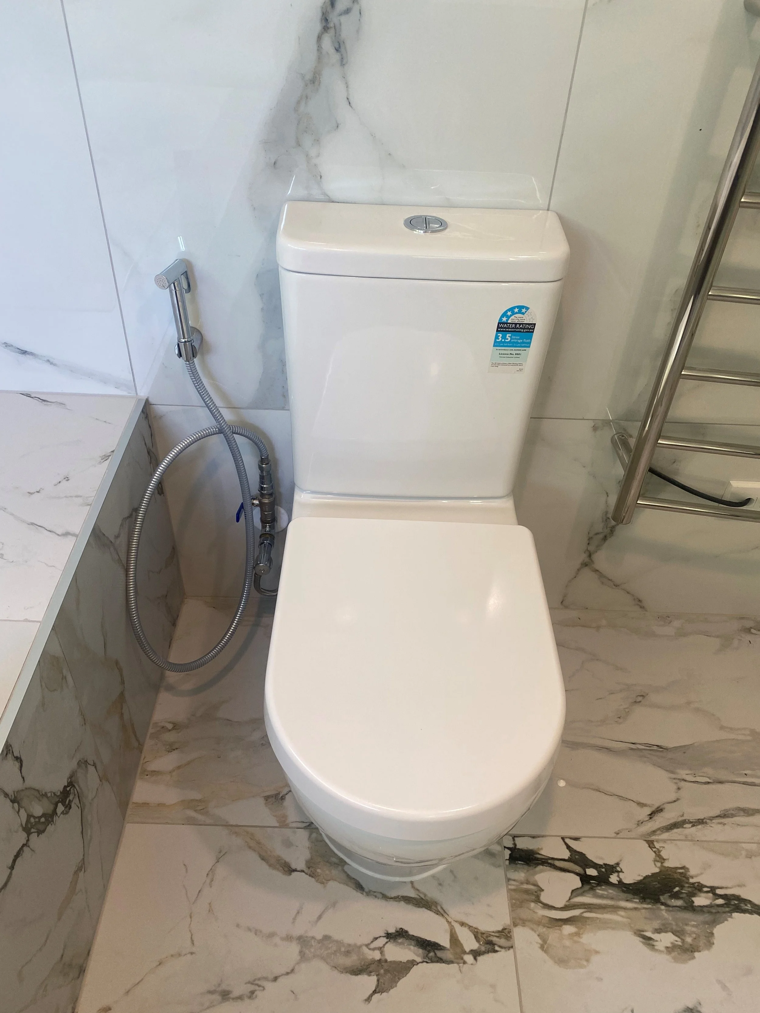 A modern bathroom toilet with a bidet spray attached on the left side, situated on marble tiled floor and wall.