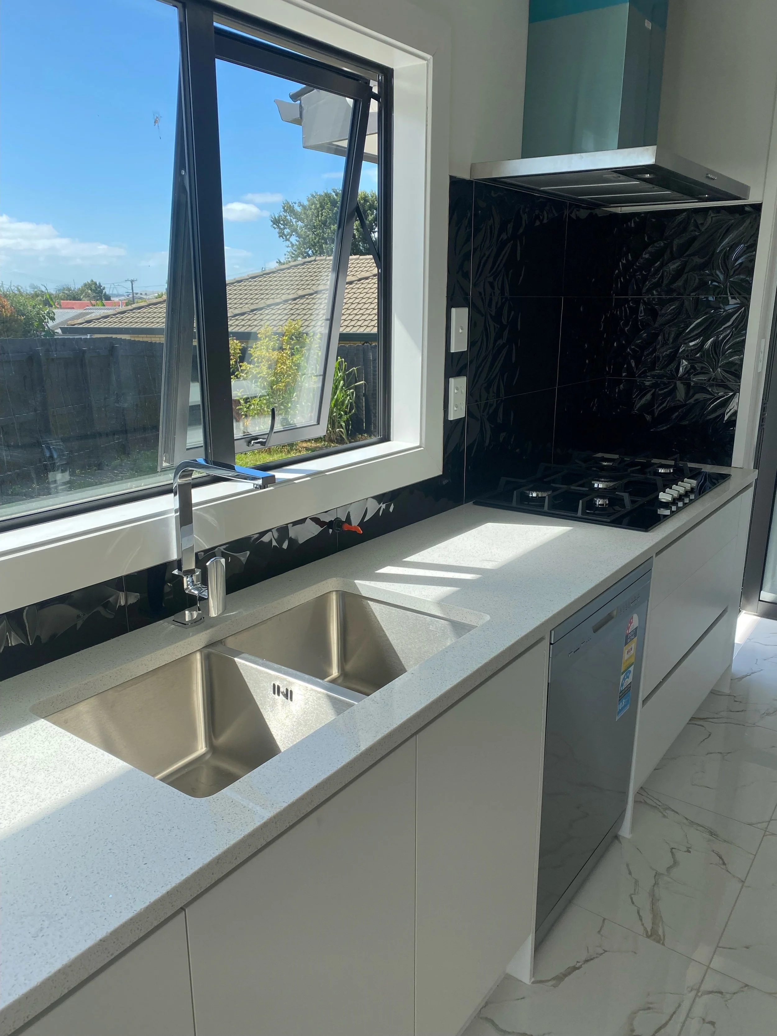 Modern kitchen with a double stainless steel sink, white countertops, black patterned backsplash, and a gas stove, with a window showing a backyard and a blue sky.