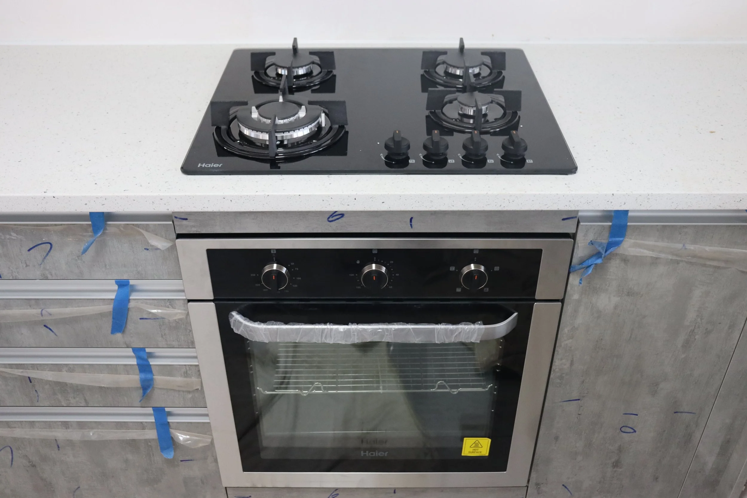 Black cooktop with four gas burners and control knobs, installed above a stainless steel oven with a glass door in a kitchen under construction.