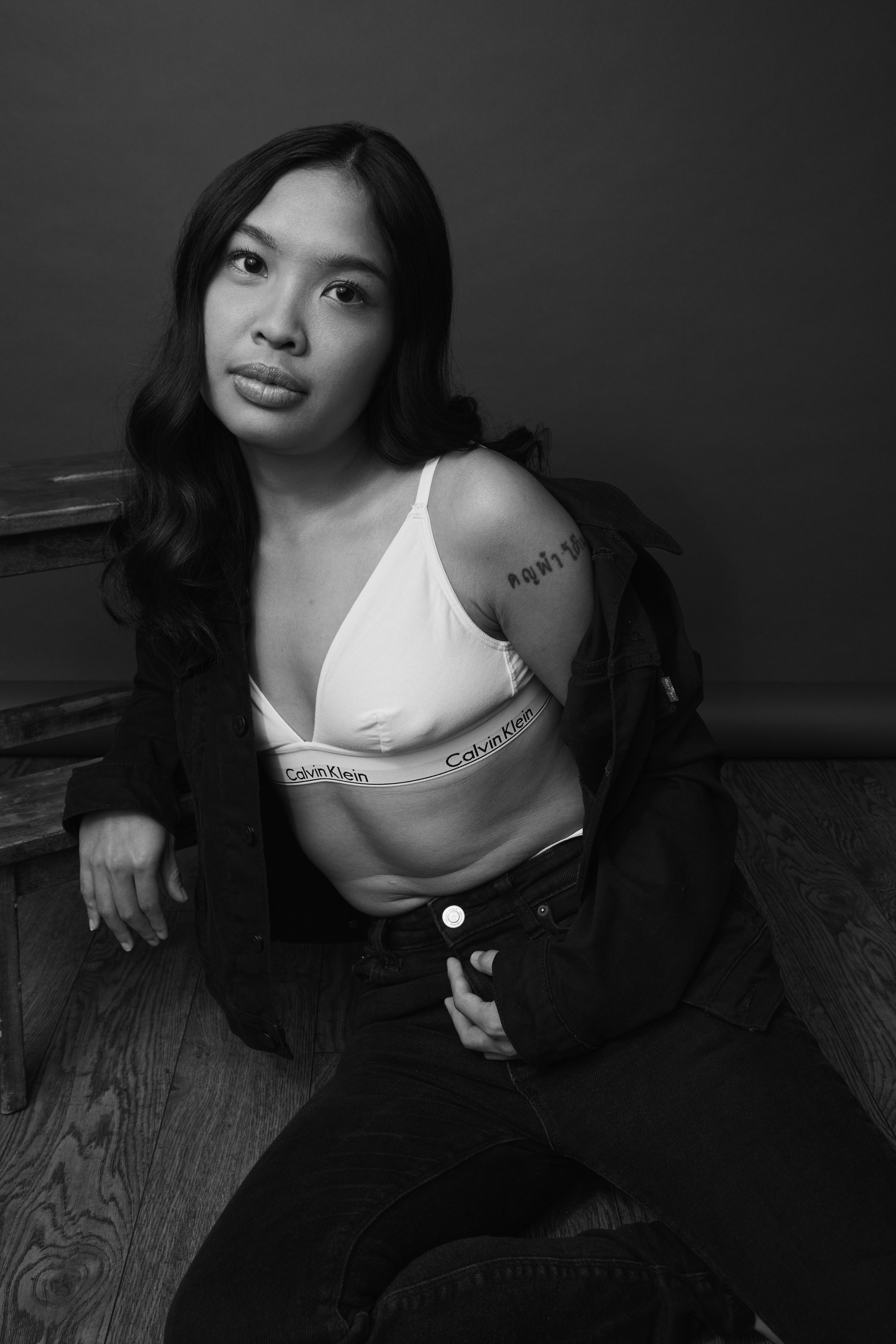 Black and white photo of a woman with long wavy hair, wearing a white Calvin Klein bra and black jeans, sitting on a wooden floor with a dark background.