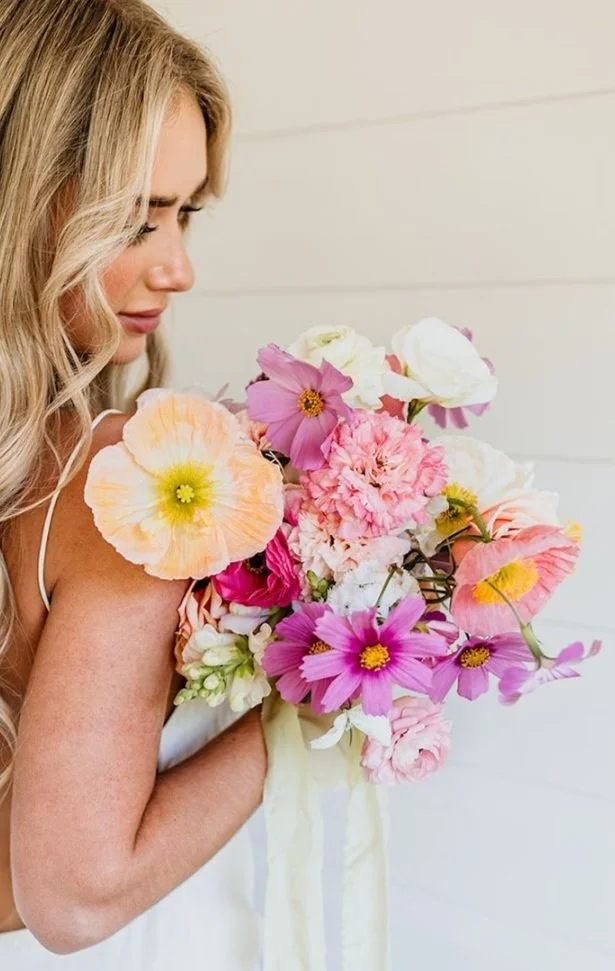 A woman with long blonde hair holding a vibrant bouquet of pink, white, peach, and purple flowers in front of a white wooden wall.