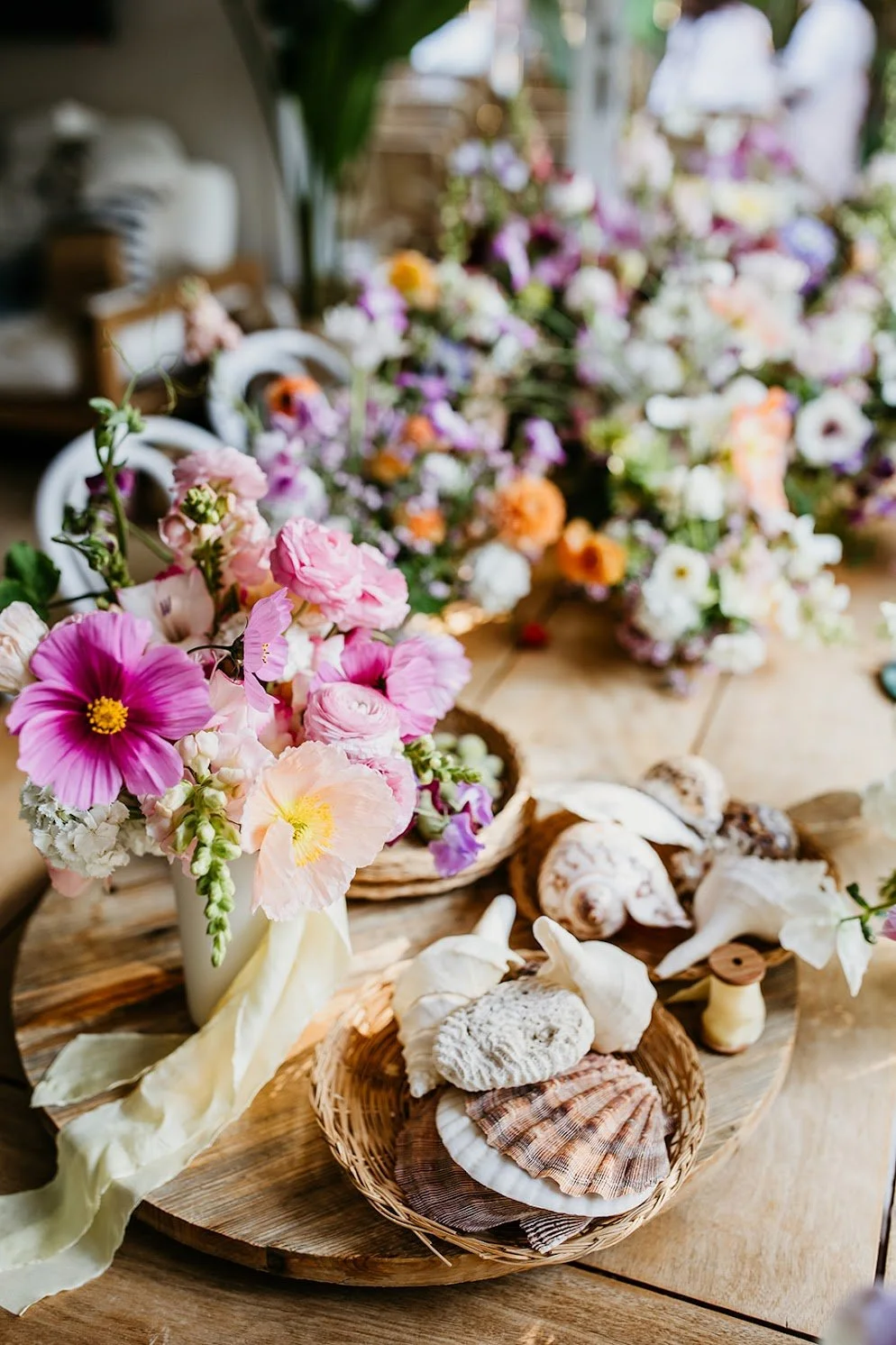 A table decorated with seashells, flowers in vases, and wooden accents, with a blurred background of plants and outdoor setting.
