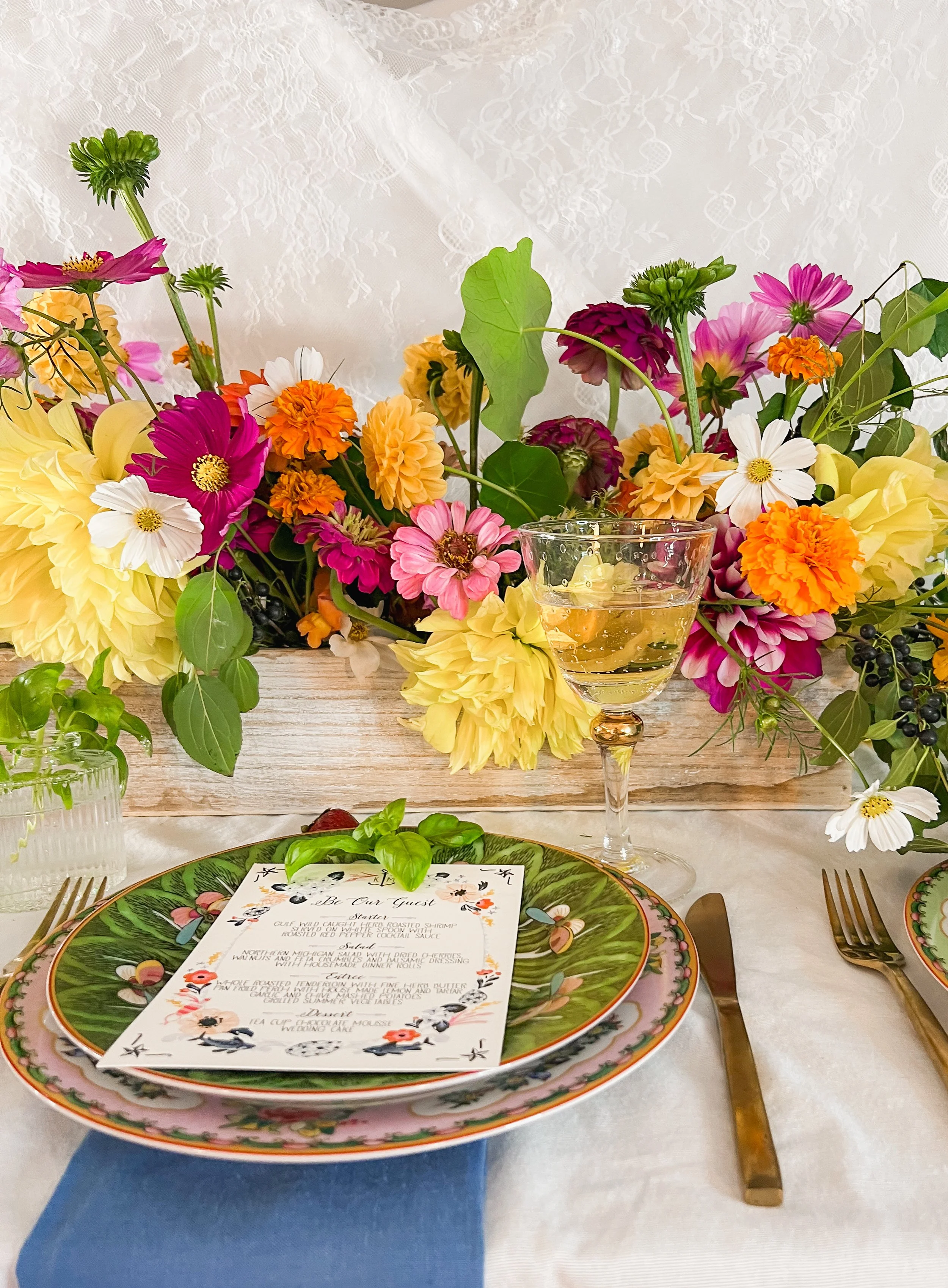 A table setting with floral plates, gold utensils, a menu, a glass of white wine, and a large colorful flower arrangement in the background.
