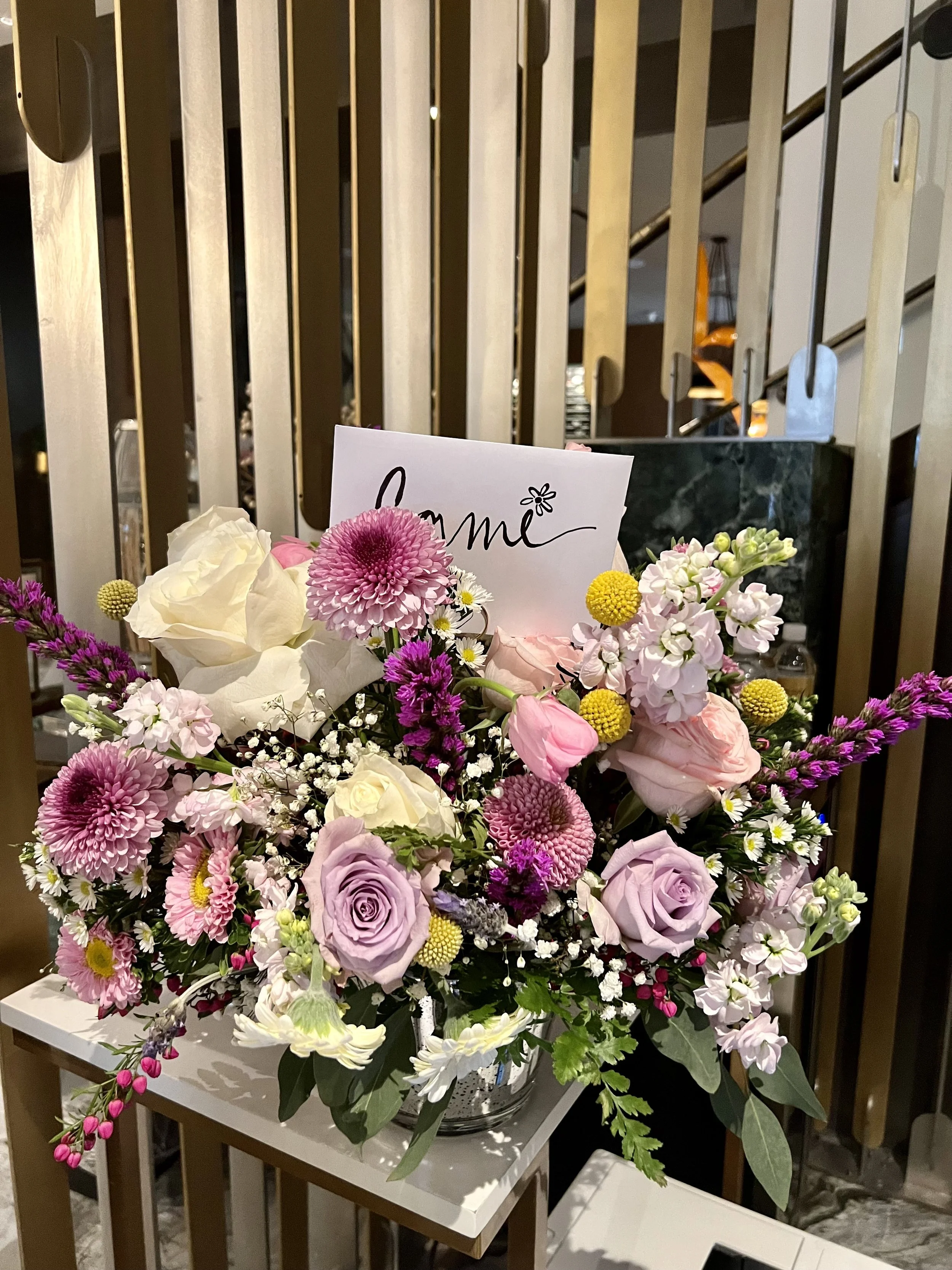 A colorful bouquet of flowers that includes pink, purple, white, and yellow blossoms with a small sign that says 'love' in cursive writing, placed on a small white table in front of wooden slats with a staircase in the background.
