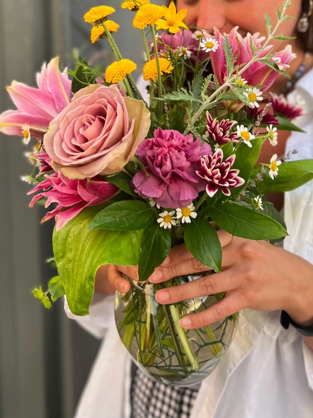 Person holding a glass vase filled with a colorful bouquet of pink roses, yellow marigolds, pink lilies, white daisies, and various green leaves.