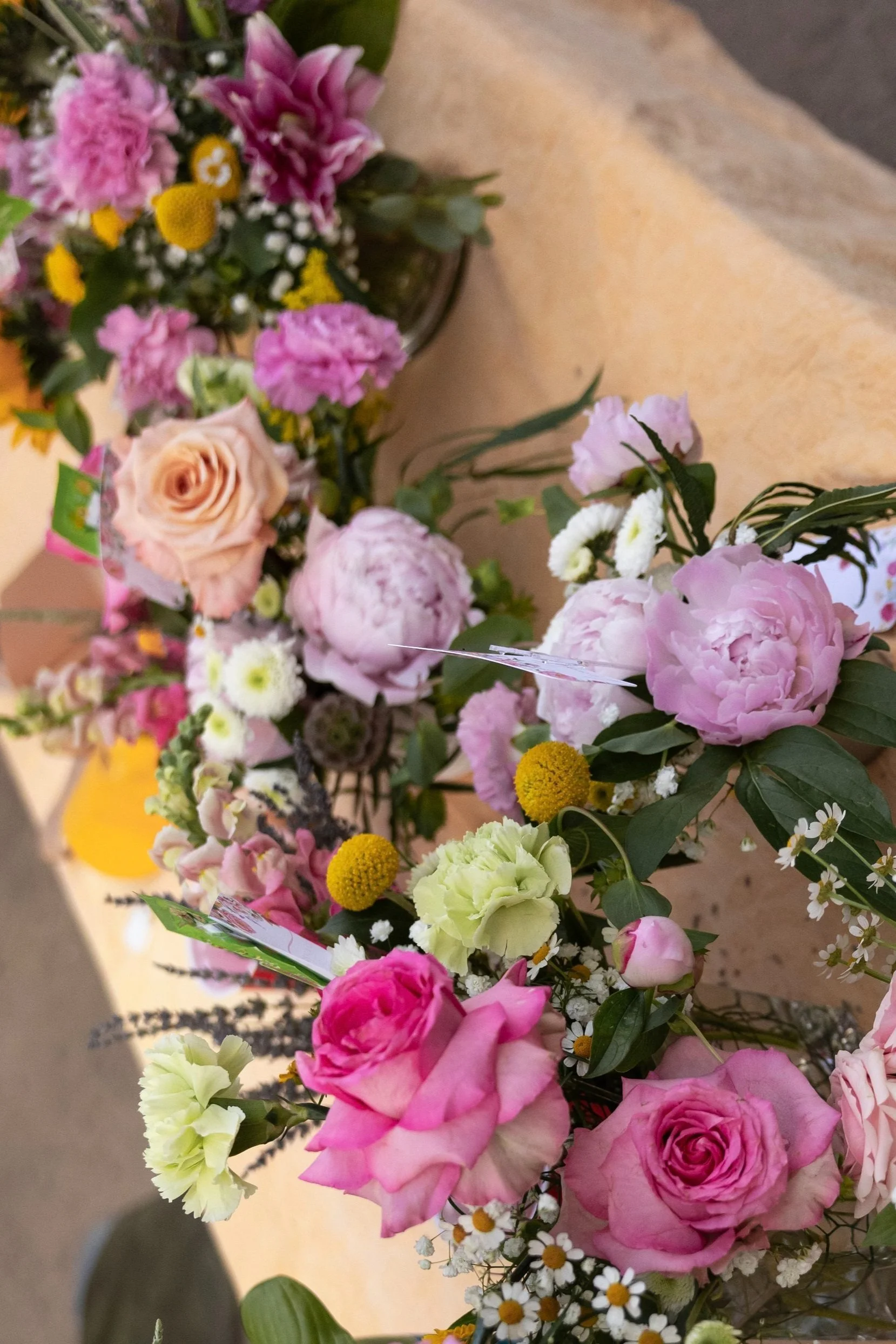 Various colorful flower bouquets with pink roses, peonies, yellow billy balls, white daisies, and other assorted flowers on a wooden surface.