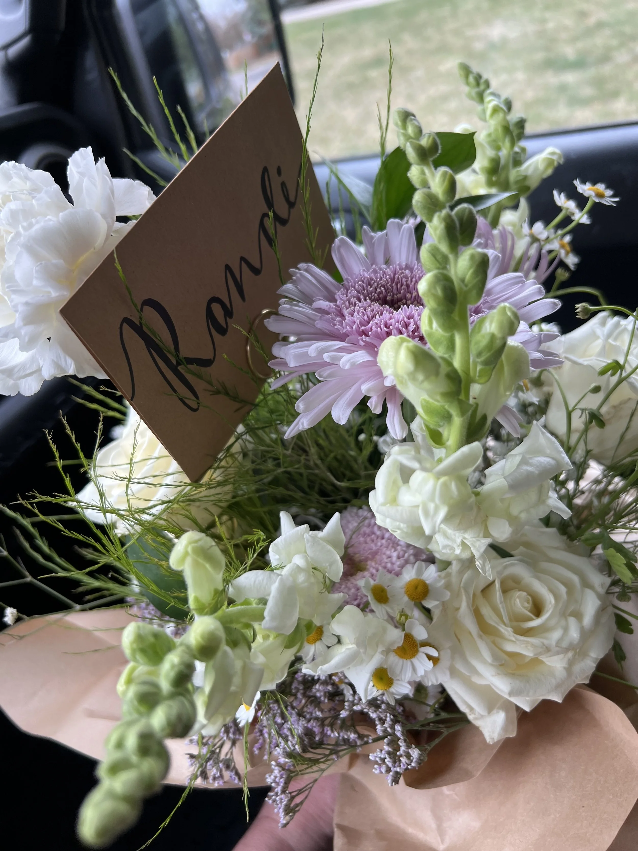A bouquet of mixed white, pink, and purple flowers with greenery, including daisies, roses, and snapdragons, with a brown paper wrapping and a brown card that says "Love" in black cursive.