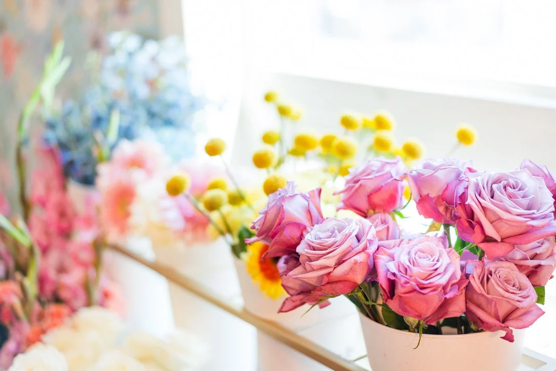 Pink roses in a white vase on a table with colorful flowers in the background