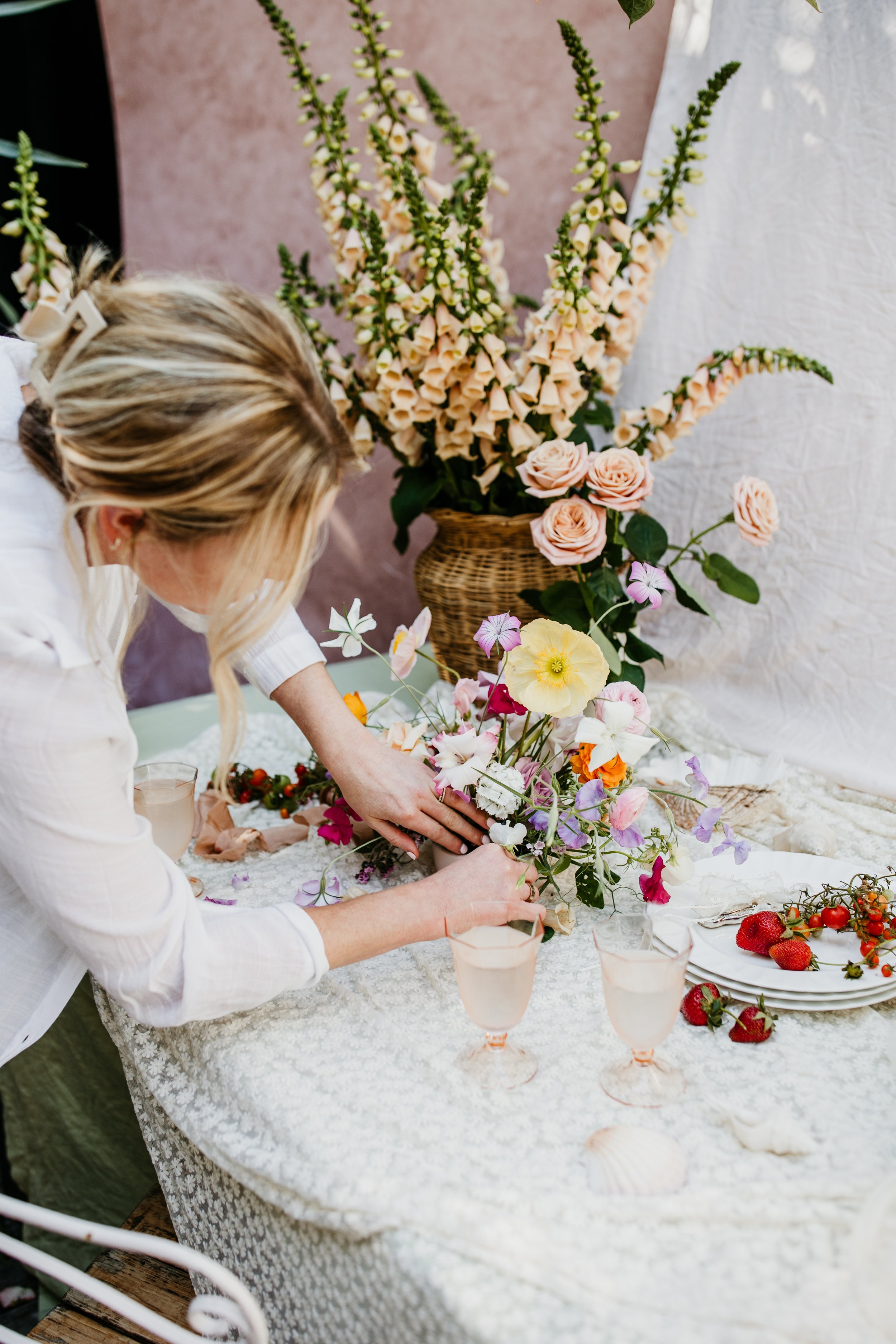 A woman arranging a floral centerpiece on a table with various flowers, strawberries, and glasses of a pink beverage, with large peach-colored snapdragons and smaller pink and purple flowers, in a setting with soft pink and white backgrounds.