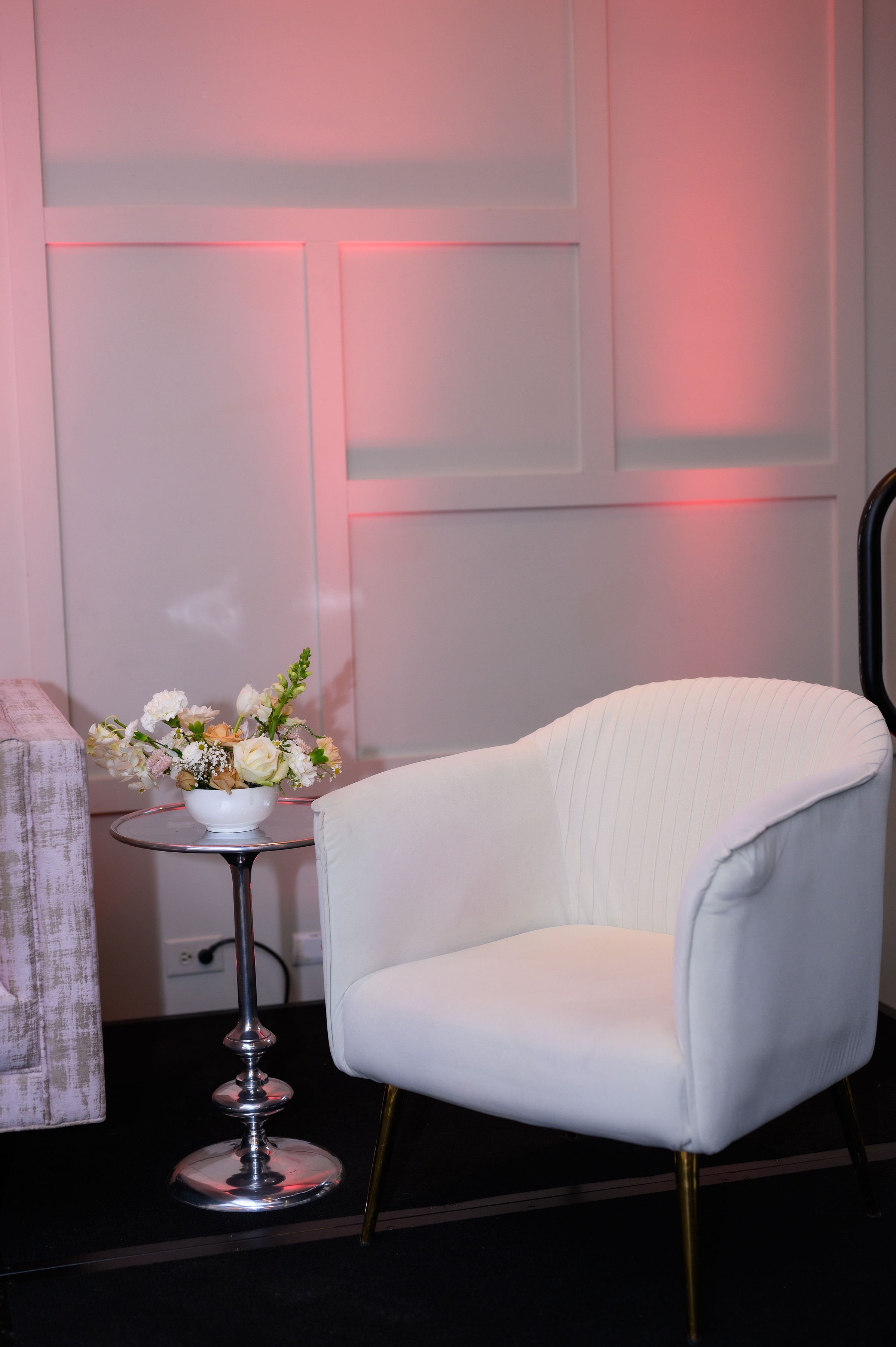 A white armchair with gold legs, a flower arrangement on a small round table, against a wall with pink lighting in the background.