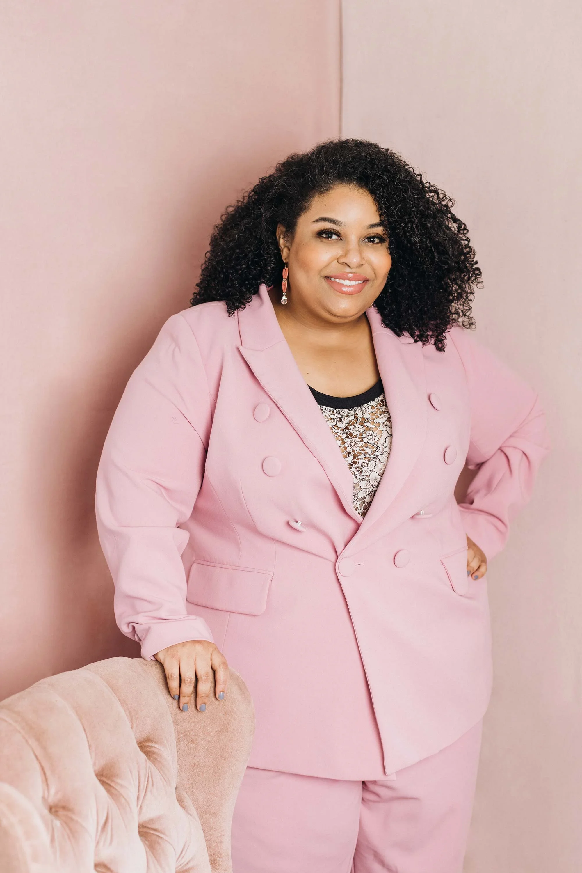 Jordan Maney wears a pink suit while standing in front of a pink wall and behind a pink chair