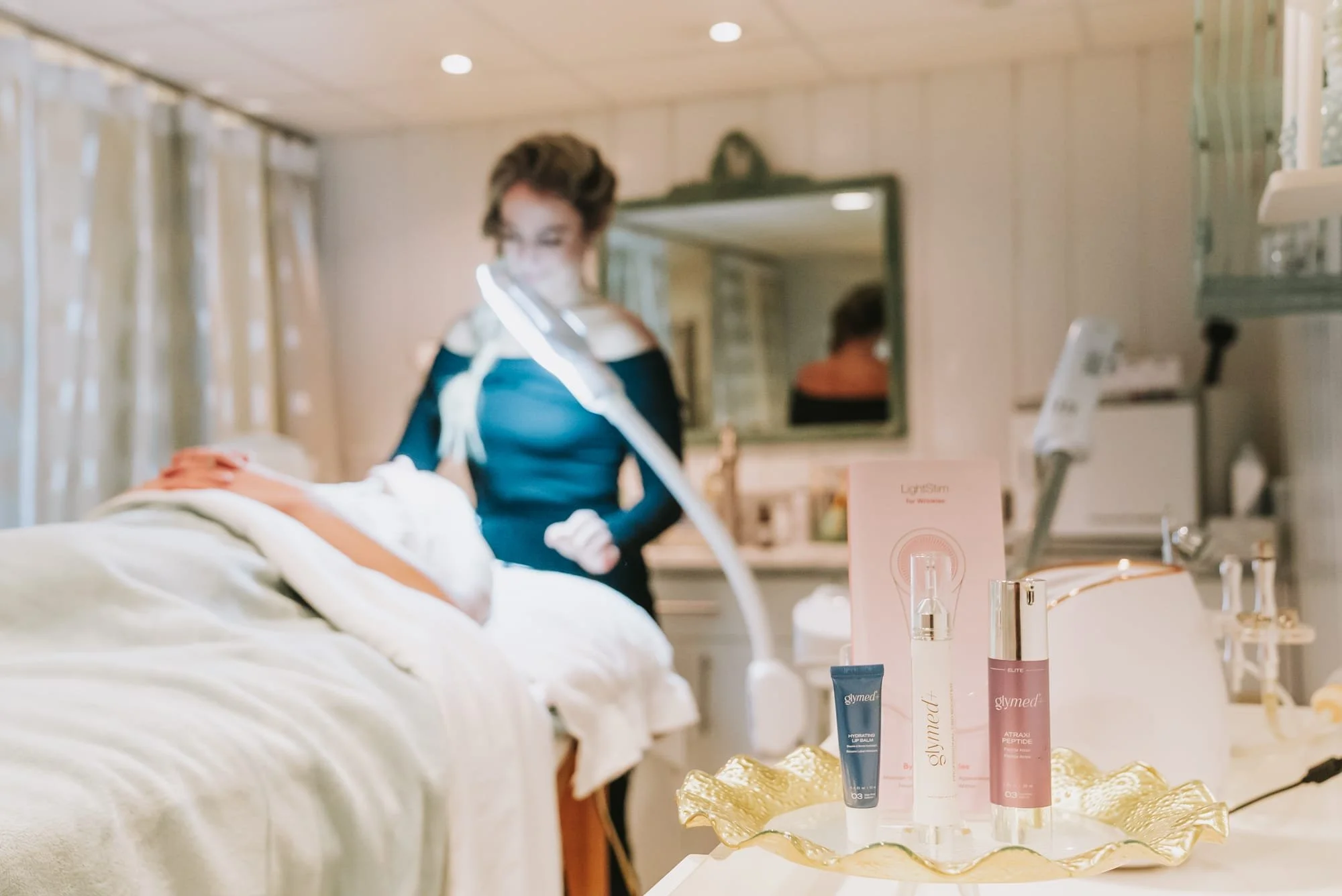 A woman receiving a facial treatment in a spa or skincare clinic, with skincare products on a golden tray in the foreground.