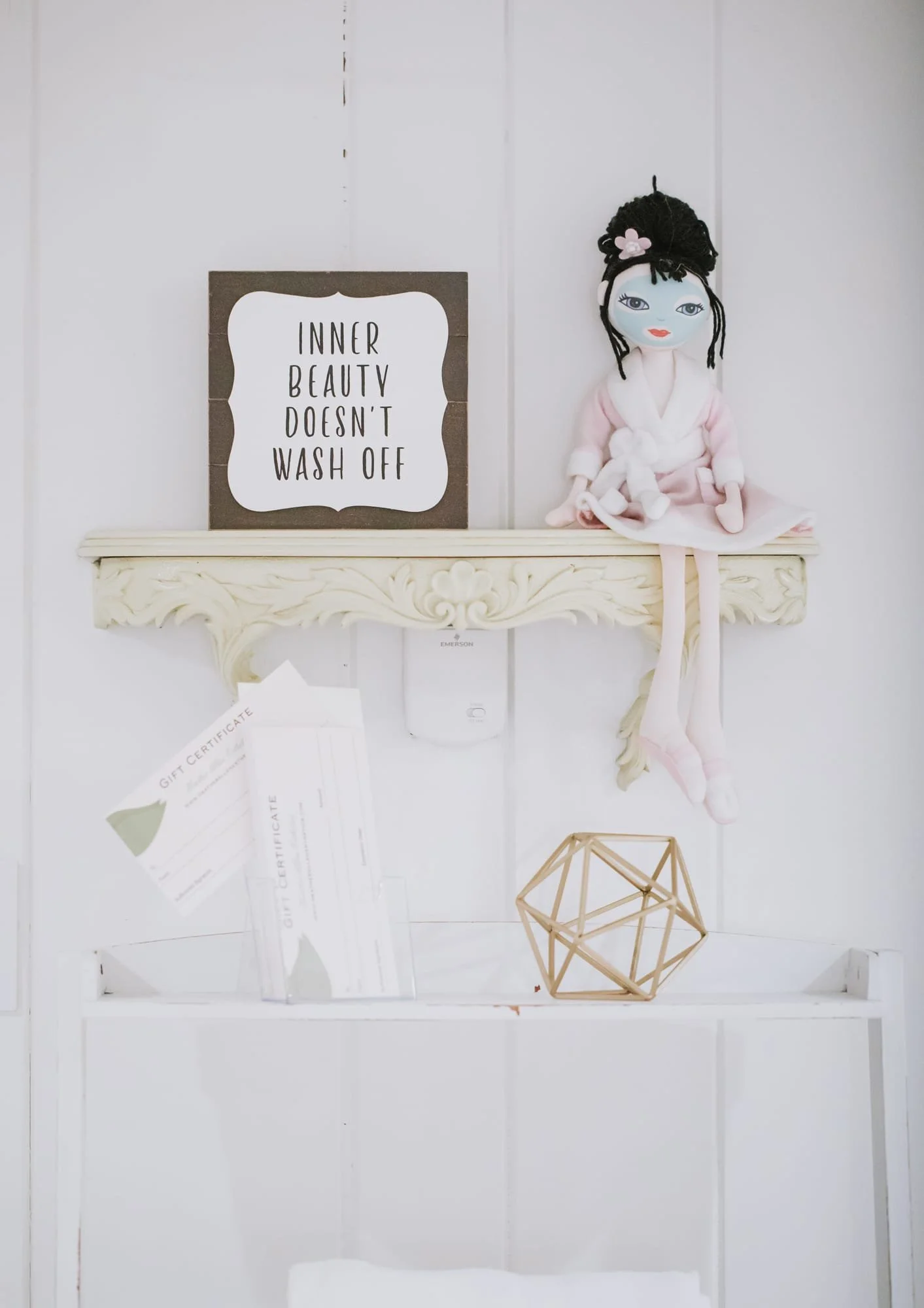 Decorative shelf with a sign saying "Inner Beauty Doesn't Wash Off," a plush doll, and some papers and a geometric sculpture underneath.